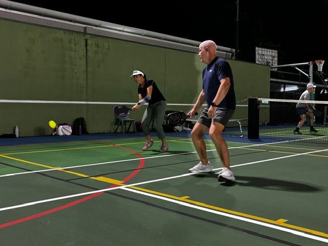Two people playing pickleball on an outdoor court at night, with a yellow ball in play.