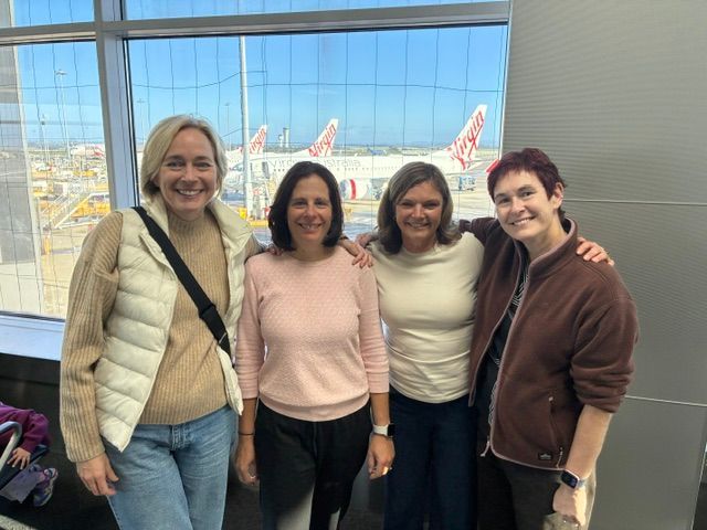 Four women smiling at an airport terminal window with planes visible outside