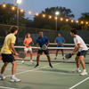 Five people play pickleball on an outdoor court at night under string lights.