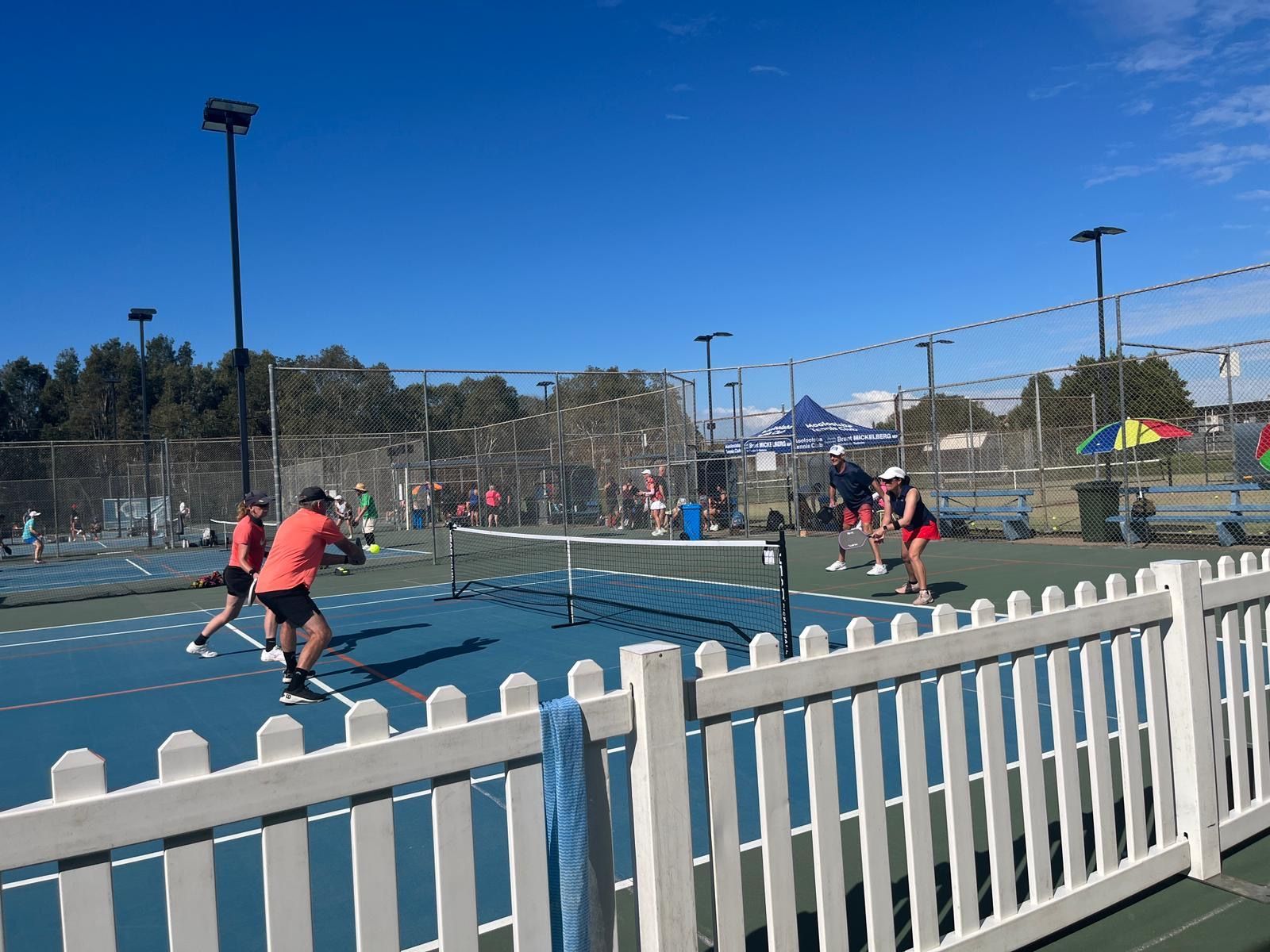 Paddle tennis players on an outdoor blue court behind a white fence on a sunny day