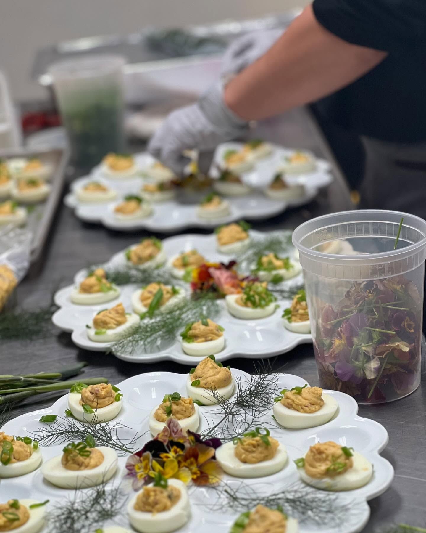 Plated deviled eggs being arranged on trays at a food prep table with gloves and a clear cup of greens