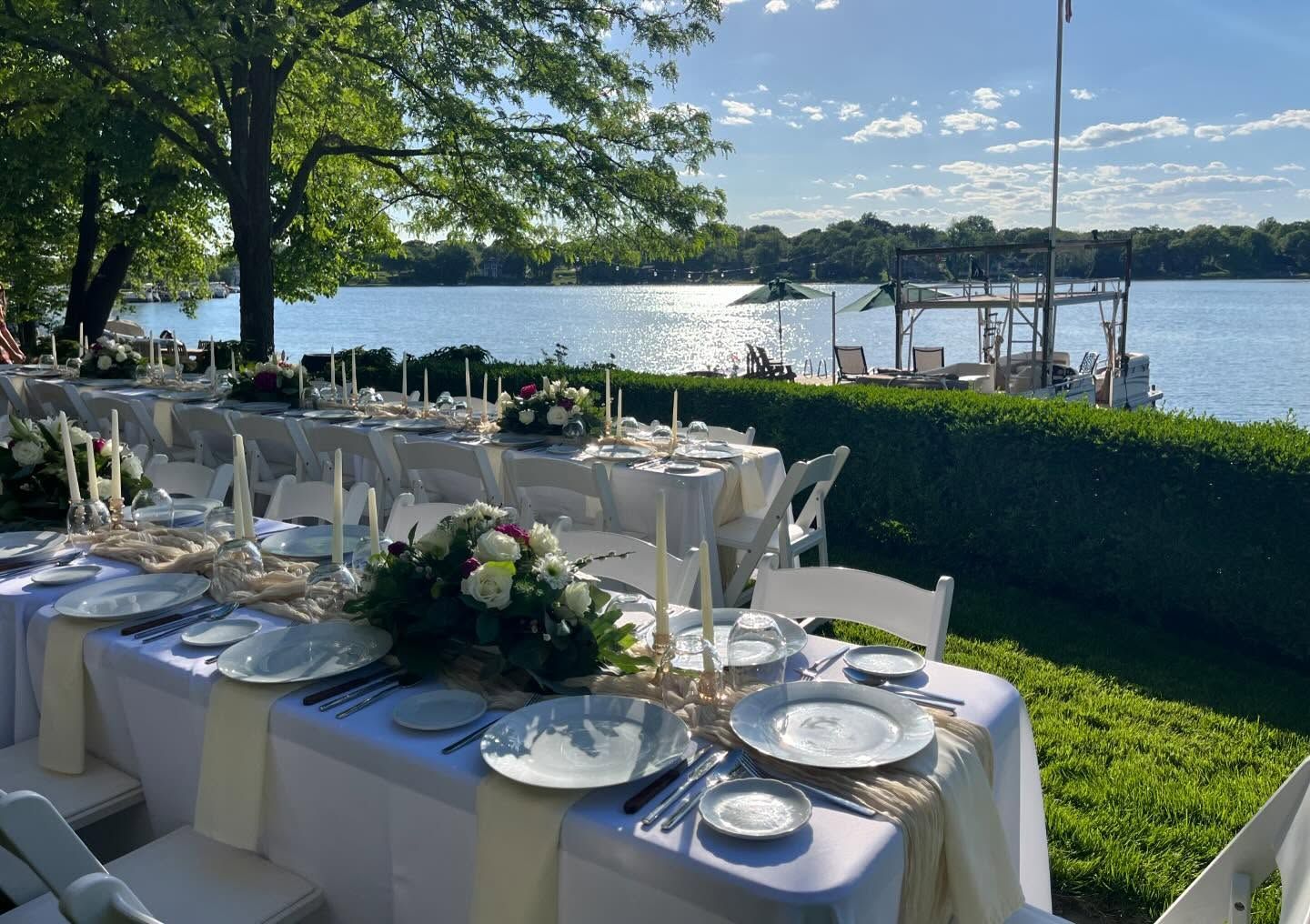 Elegant outdoor dining setup on a lakeside lawn with white tables, flowers, and views of the water