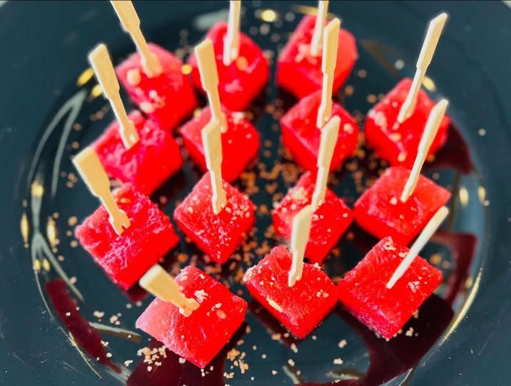 Red cube desserts on toothpicks arranged on a dark plate with scattered crumbs.