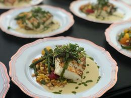 Plated gourmet fish with grains and herbs on a white dish, with more similar plates blurred in the background