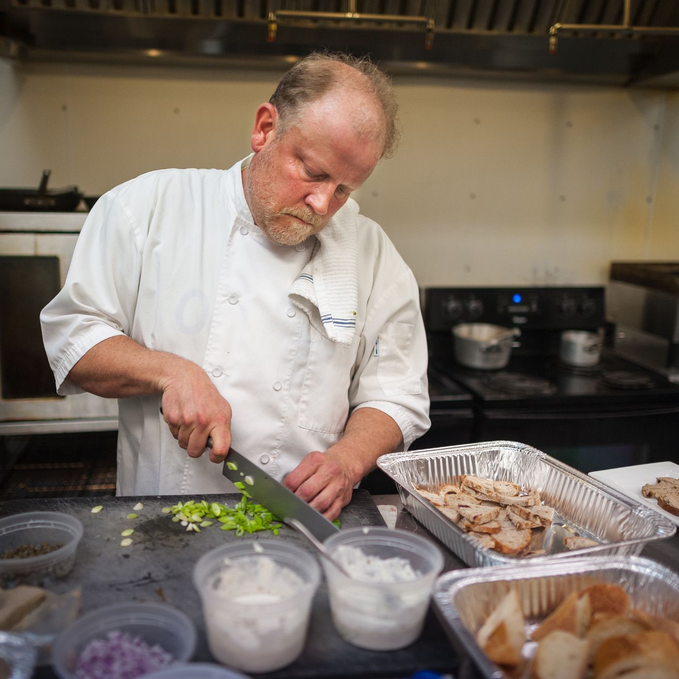 Chef chopping green onions in a kitchen beside trays of prepared food