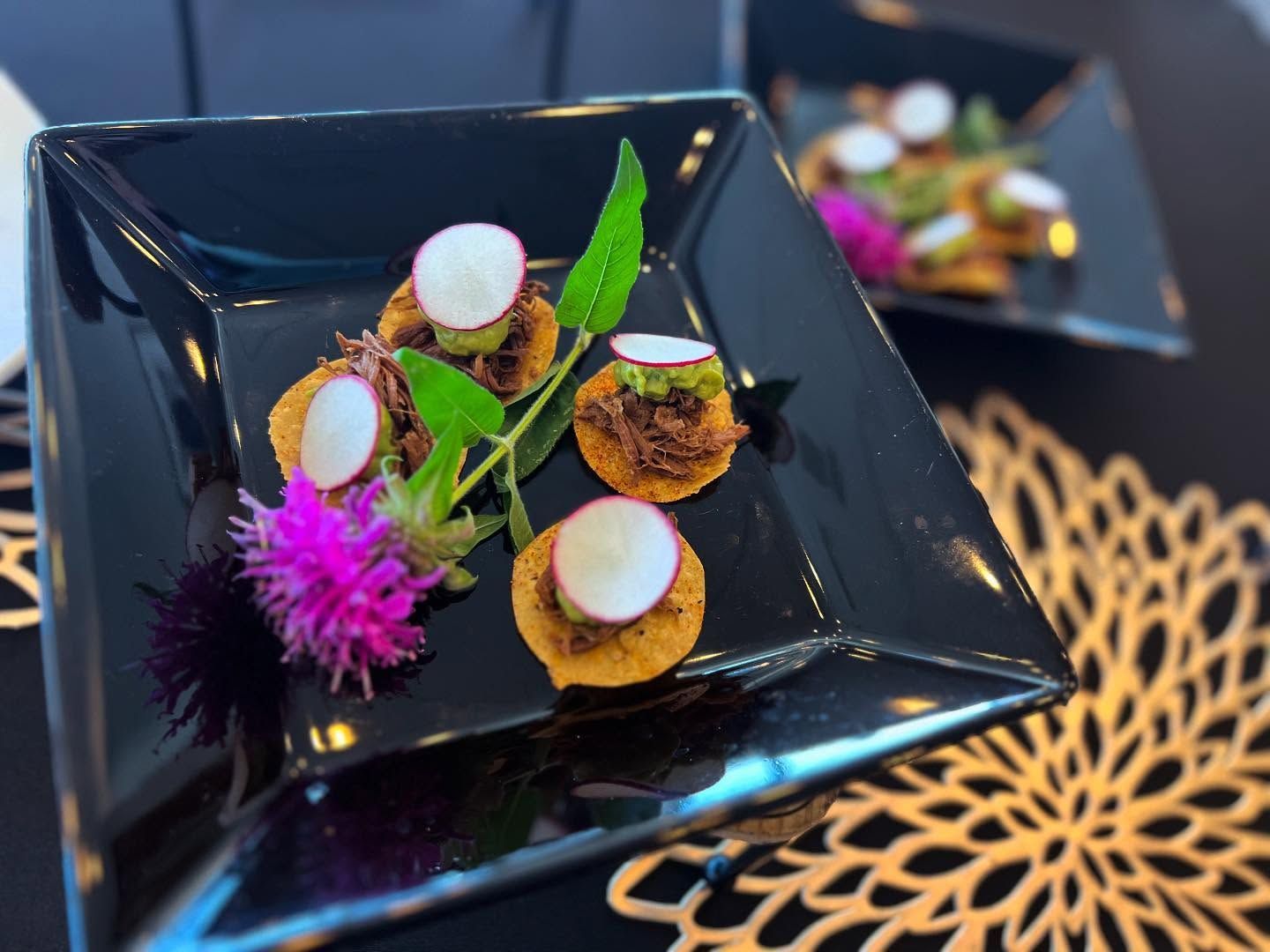 Black square plate with elegant bite-sized hors d'oeuvres, garnished with herbs and purple flowers