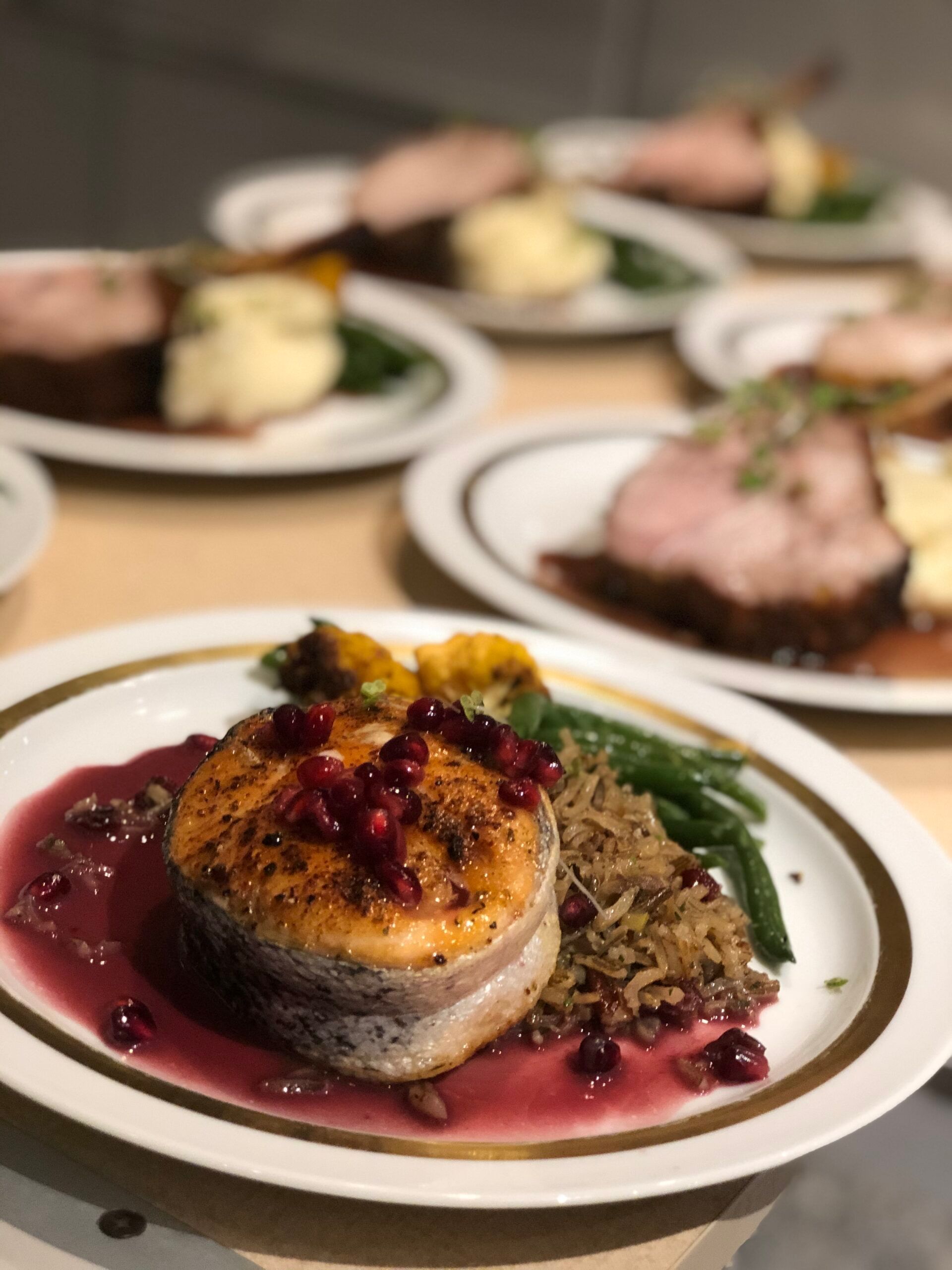 Plated gourmet meal with seared fish, red sauce, greens, and blurred dishes in the background