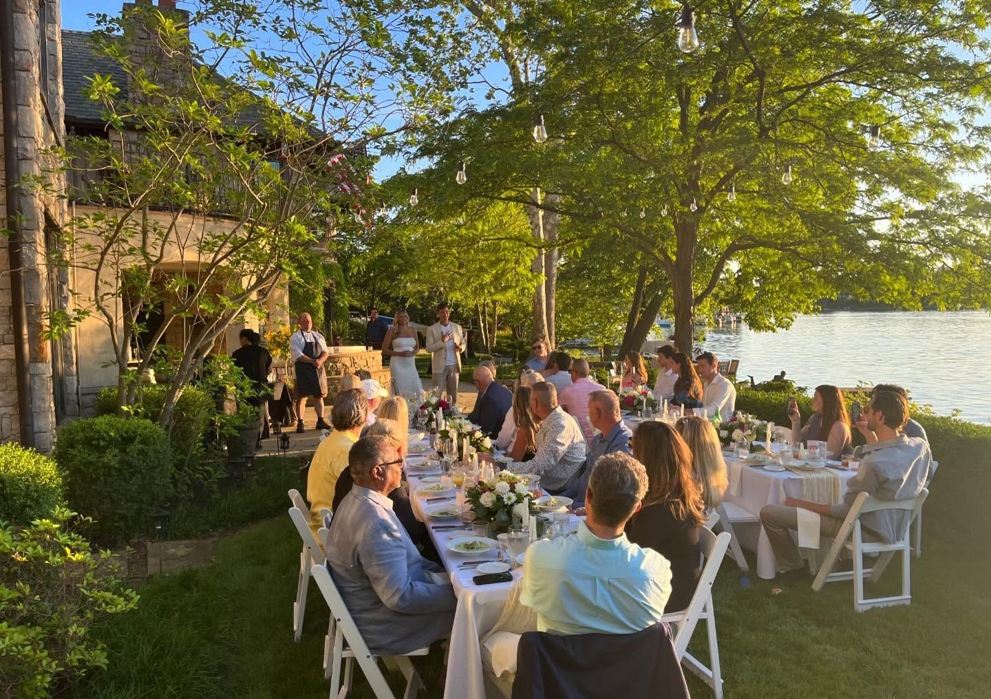 Outdoor garden dinner party with people seated at white tables under trees at sunset