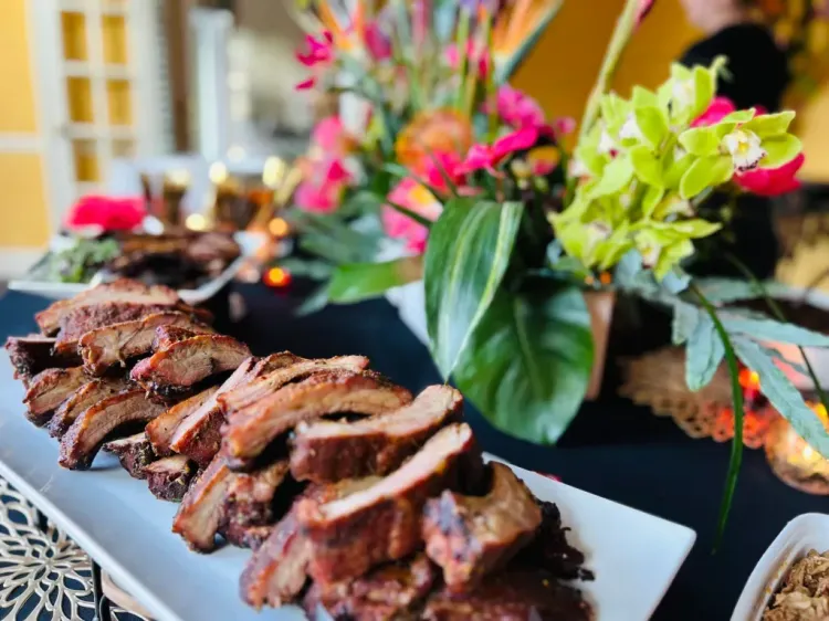Sliced grilled steak on a platter beside colorful floral centerpieces at a banquet table
