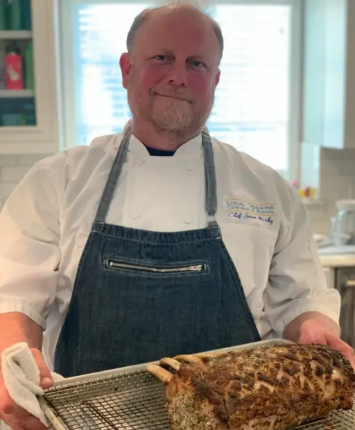 Chef in white coat and denim apron holding a tray of roasted meat in a bright kitchen