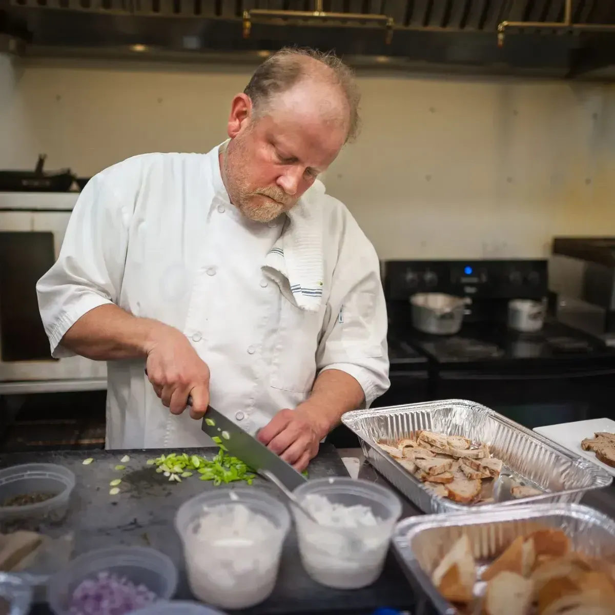 Chef chopping green onions in a kitchen beside trays of prepared food