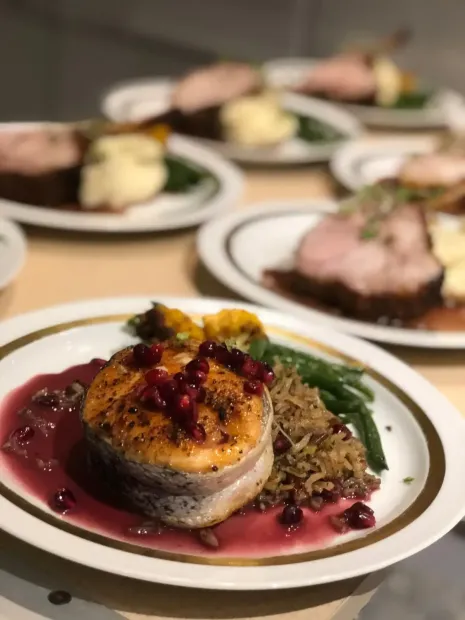 Plated gourmet meal with seared fish, red sauce, greens, and blurred dishes in the background