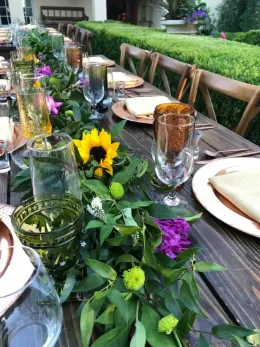 Outdoor dining table set with sunflower and purple floral centerpiece, glassware, plates, and greenery