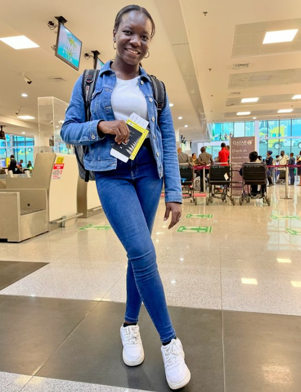 A woman in a denim jacket and jeans is standing in an airport holding a book.
