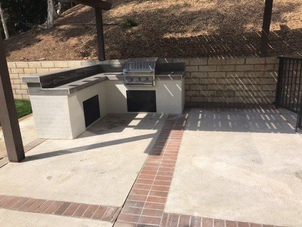 Outdoor built-in barbecue with gray and tan brick, concrete patio, and sloped hillside.