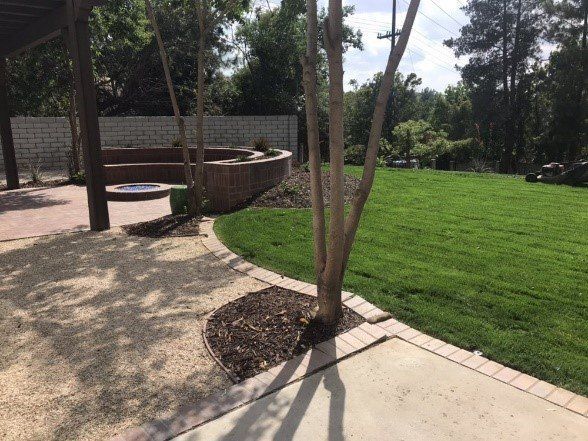 Backyard with a gravel patio, brick border, and lush green lawn, trees, and a decorative brick wall.