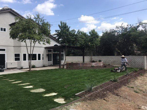 Man mowing a lush green lawn next to a two-story house, a stone pathway, and a brick wall under a partly cloudy sky.