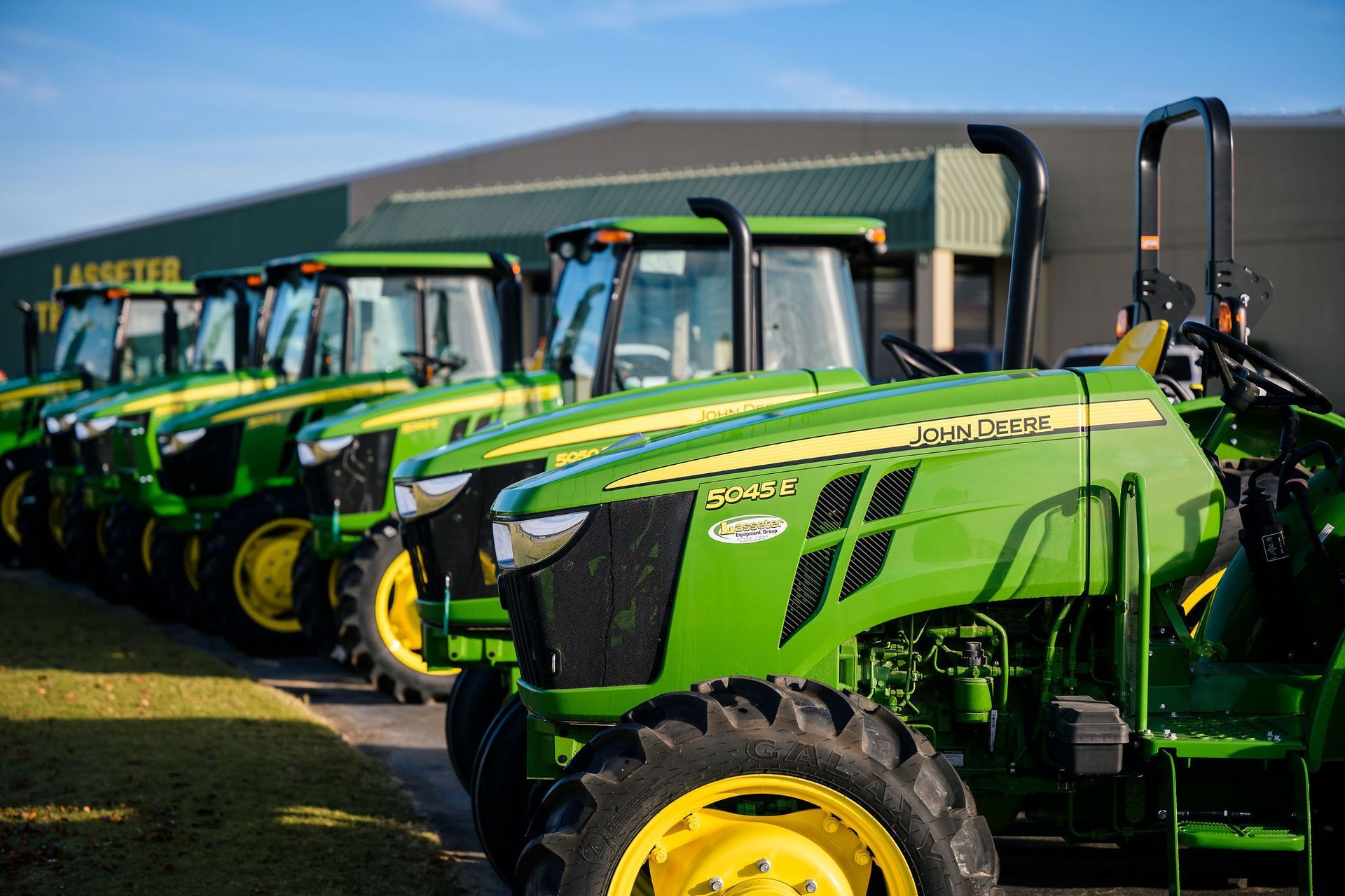 A row of green and yellow john deere tractors and a 5045e tractor are parked in front of a building.