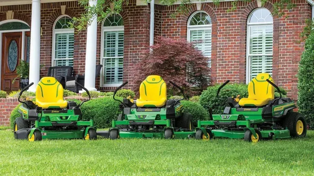 Four john deere zero turn mowers are parked in front of a brick house.