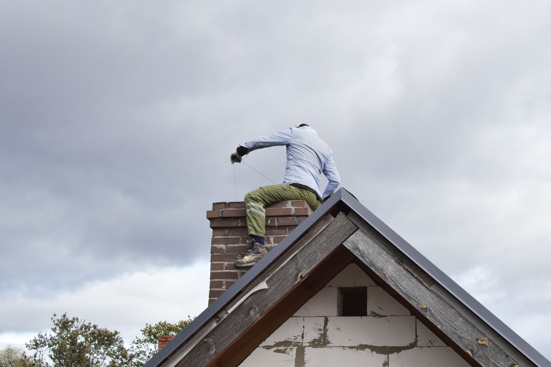 A man is sitting on top of a brick chimney on the roof of a house.