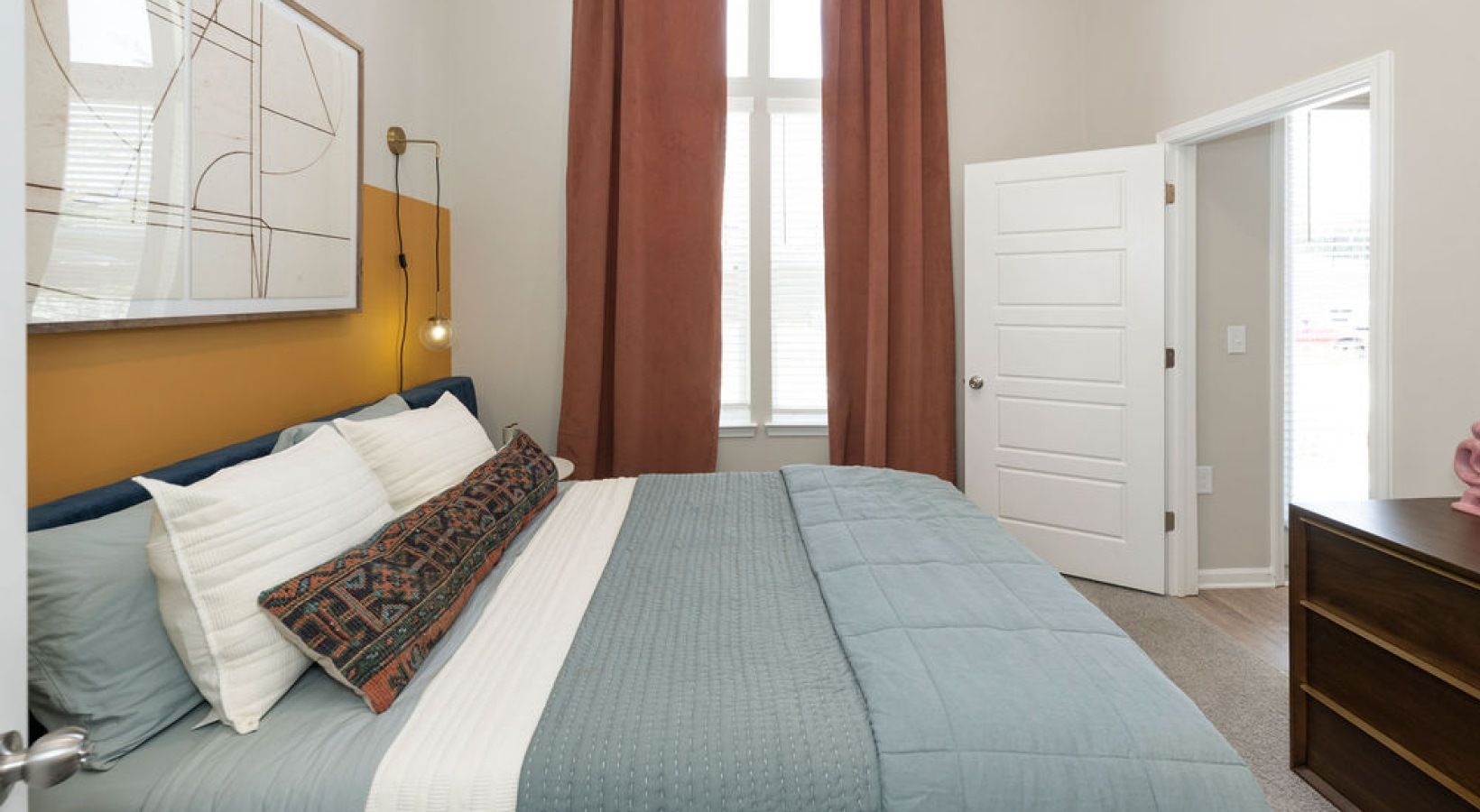 Bedroom in an apartment with blue bedding, brown curtains, and a dresser.