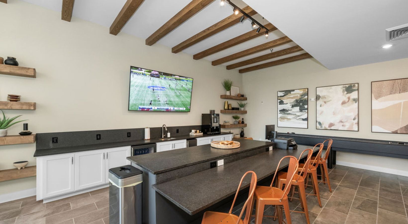 Clubhouse kitchen and lounge with an island, orange stools, exposed wooden ceiling beams, and a wall-mounted TV.