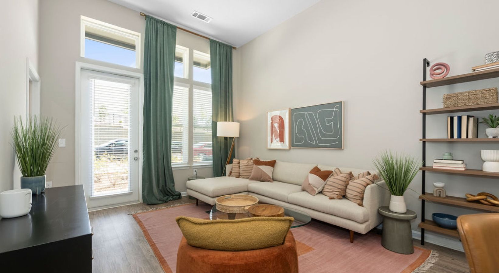 Bright apartment living room with a white sectional sofa, tall windows with green curtains, and a bookshelf.