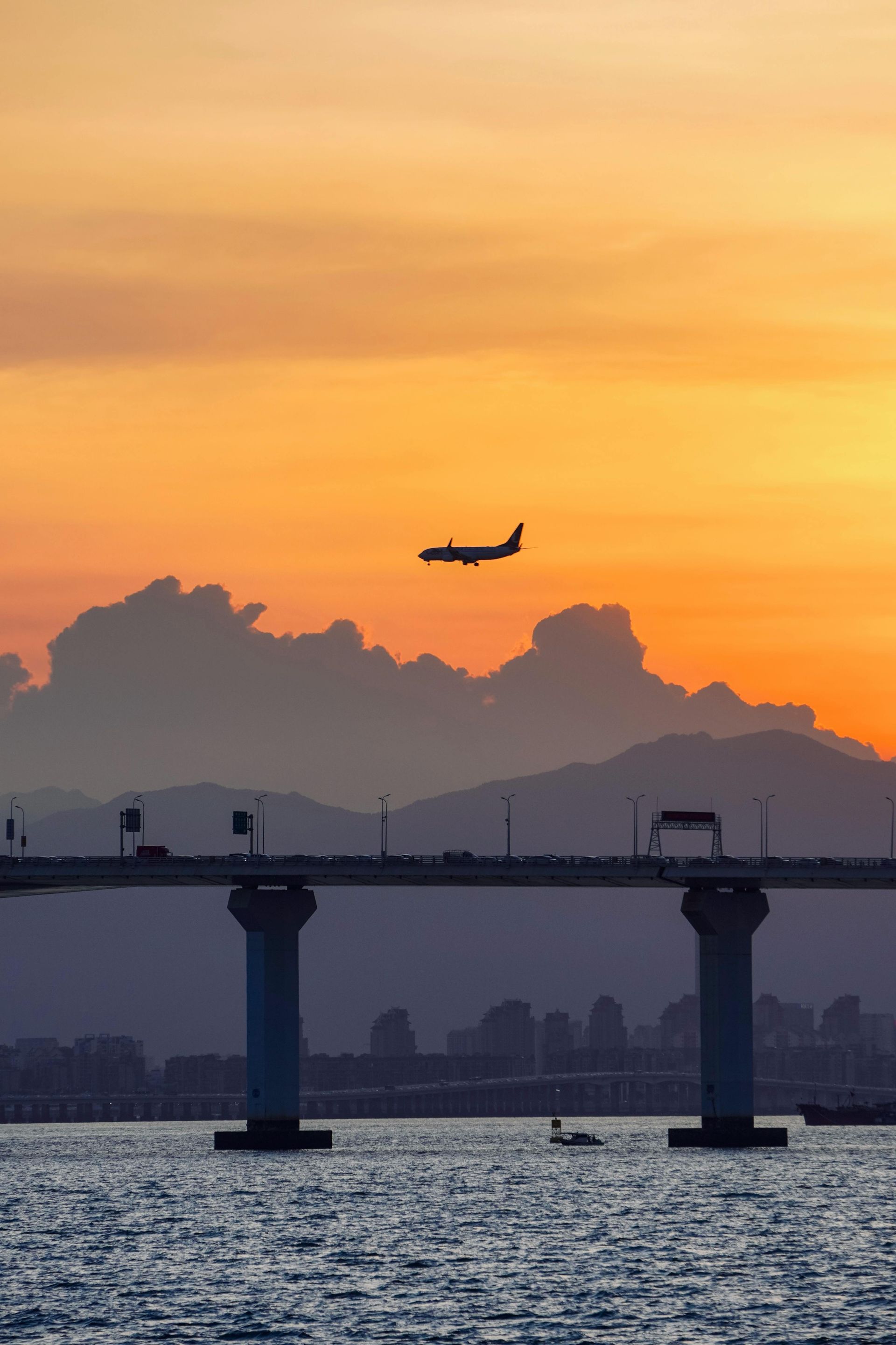 Sunset over a bridge and water, plane flying overhead. Silhouetted cars on the bridge, orange and blue hues.