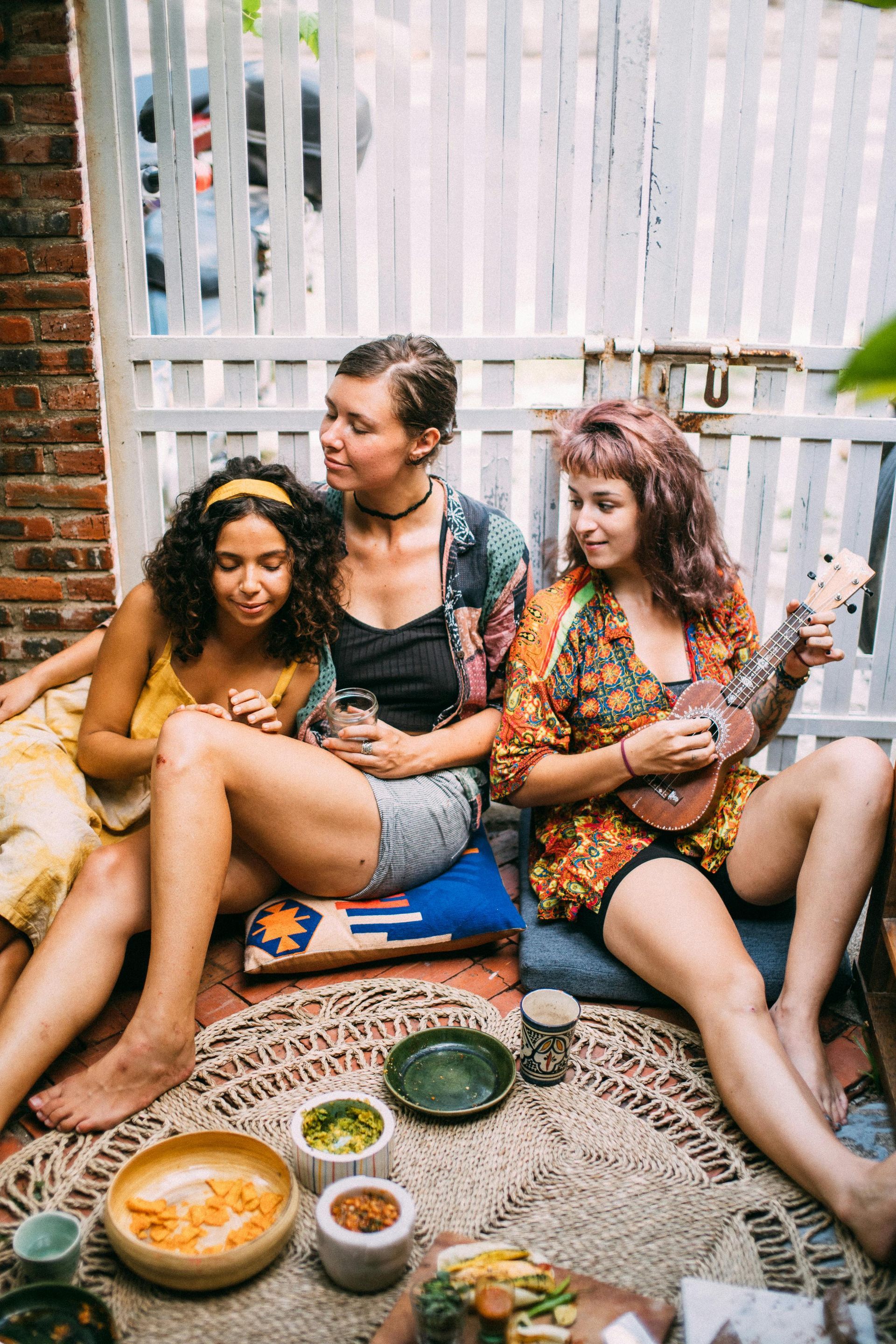 Three people seated outdoors, one plays a ukulele. They relax near food, a white fence behind them.