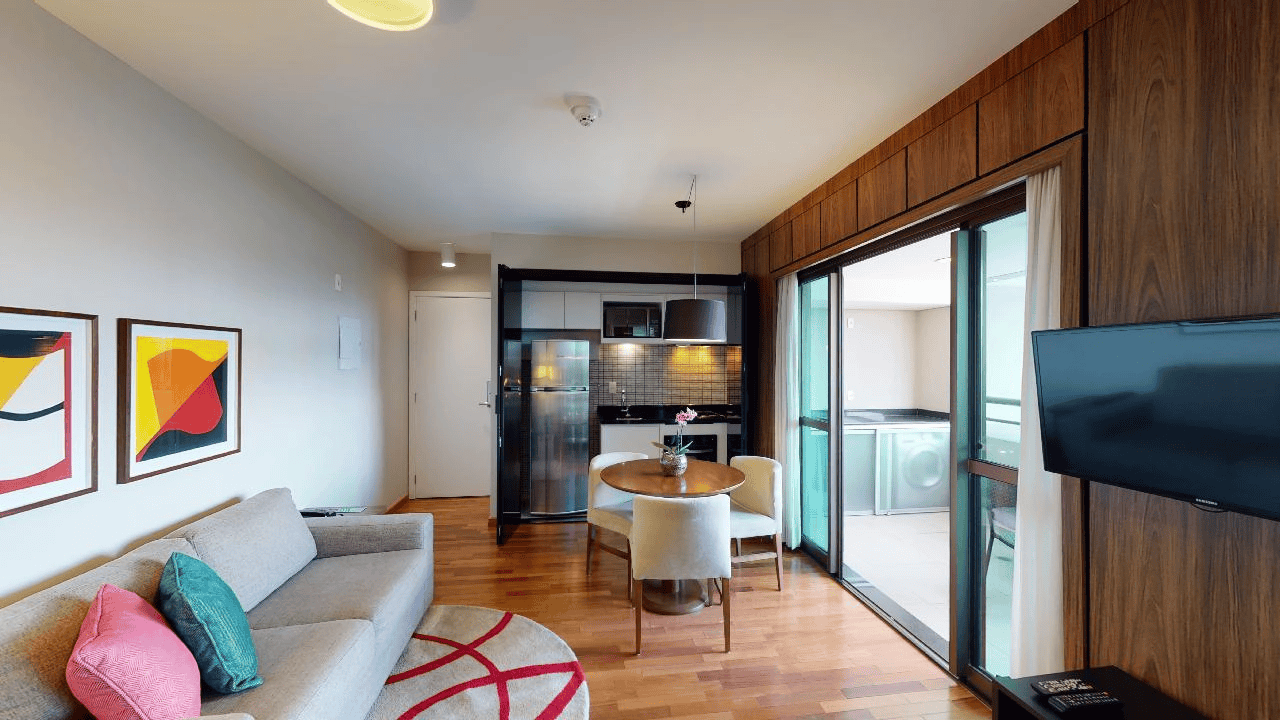 Sala de estar moderna de apartamento com sofá, mesa de jantar, cozinha compacta e portas de vidro deslizantes para varanda.