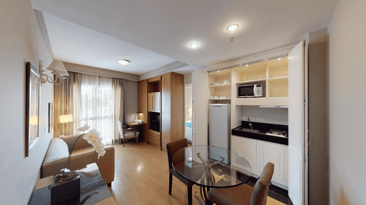 Quarto pequeno estilo hotel com cama, escrivaninha, cadeira, TV e janela com cortinas translúcidas.