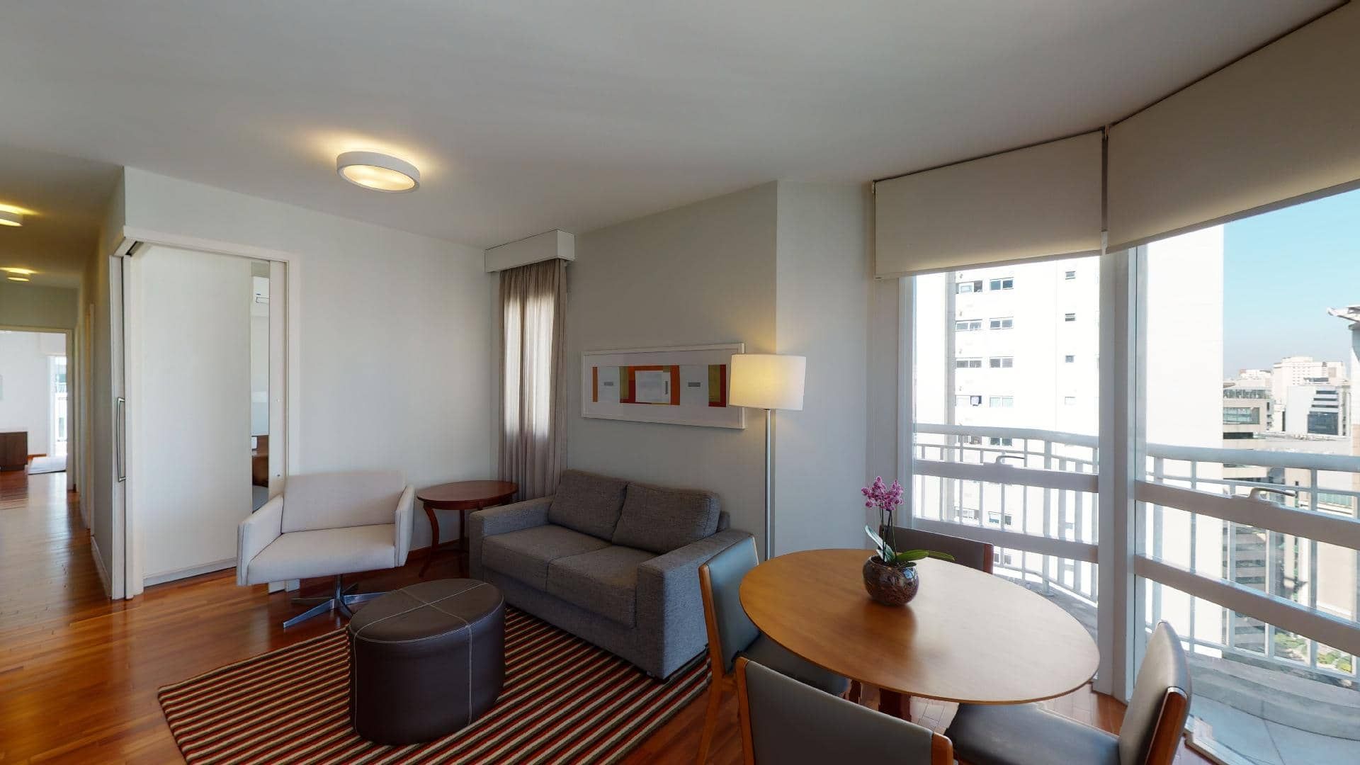 Sala de estar iluminada com sofá cinza, poltrona, mesa redonda, tapete listrado e vista da cidade através de janelas curvas.