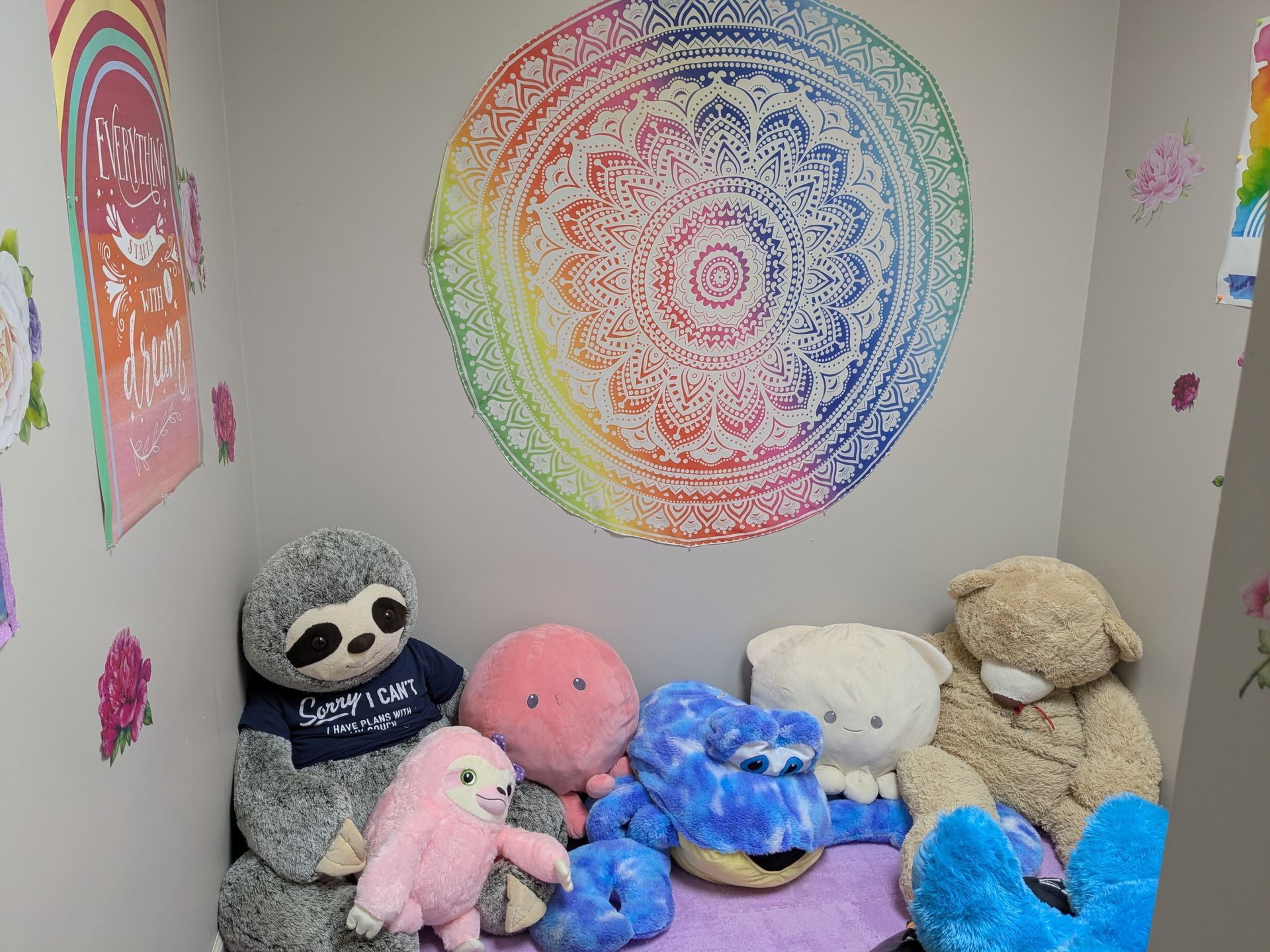 Stuffed animals in a nook with a rainbow mandala on the wall. The space is inviting with soft colors.