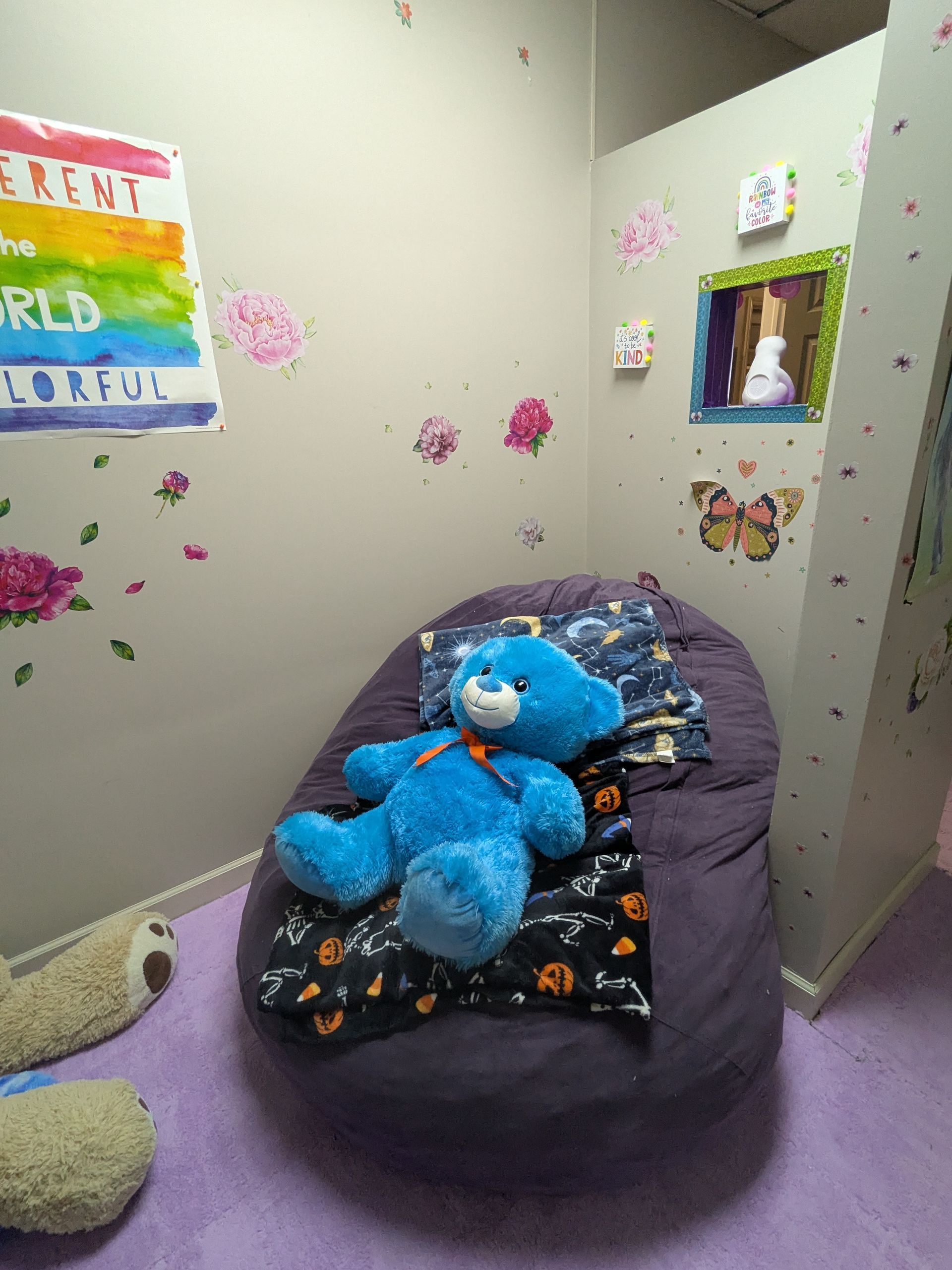 A blue teddy bear on a purple beanbag in a colorful room, decorated with flowers and a sign.