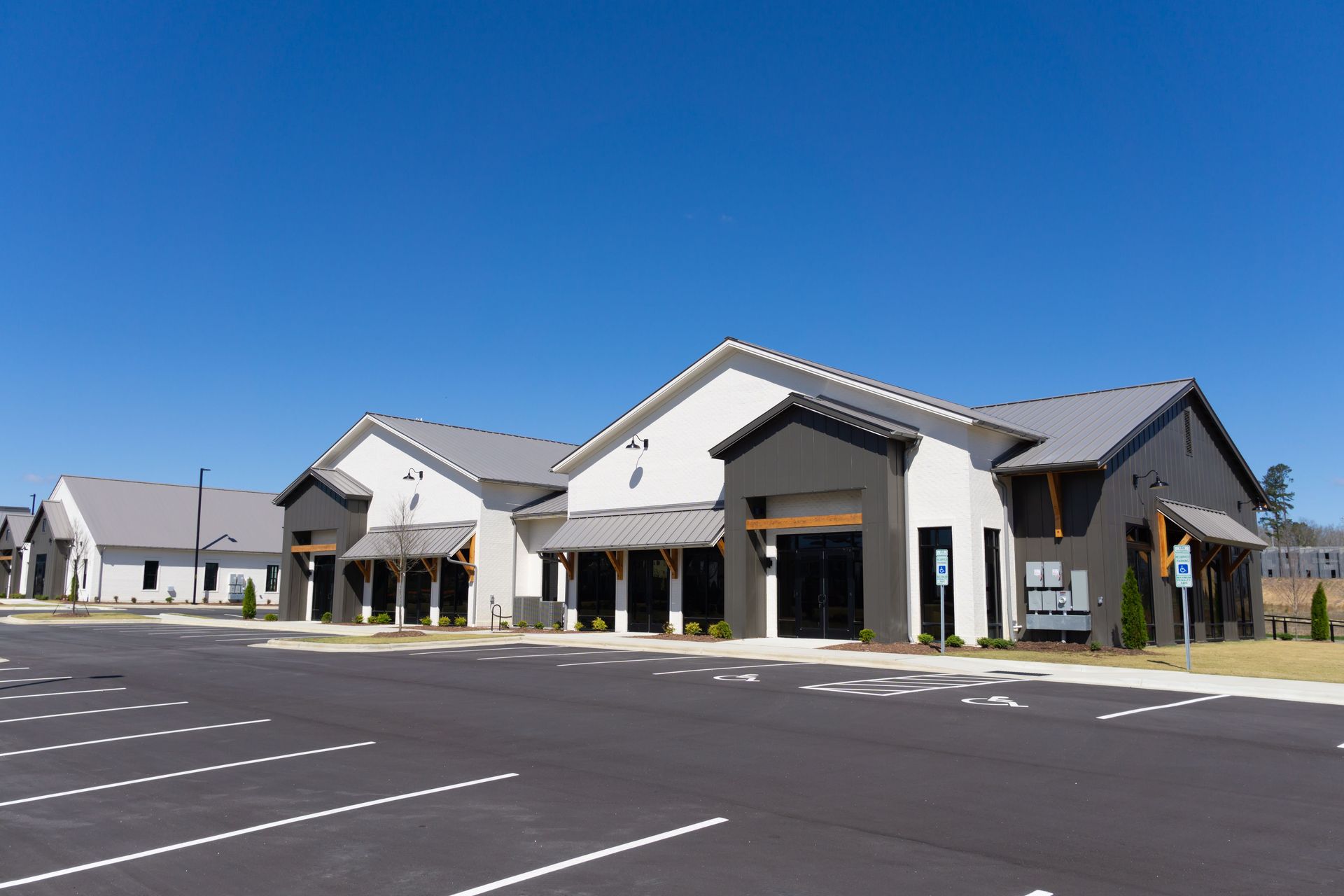 Modern, light-colored commercial building with dark gray accents and a parking lot on a sunny day.