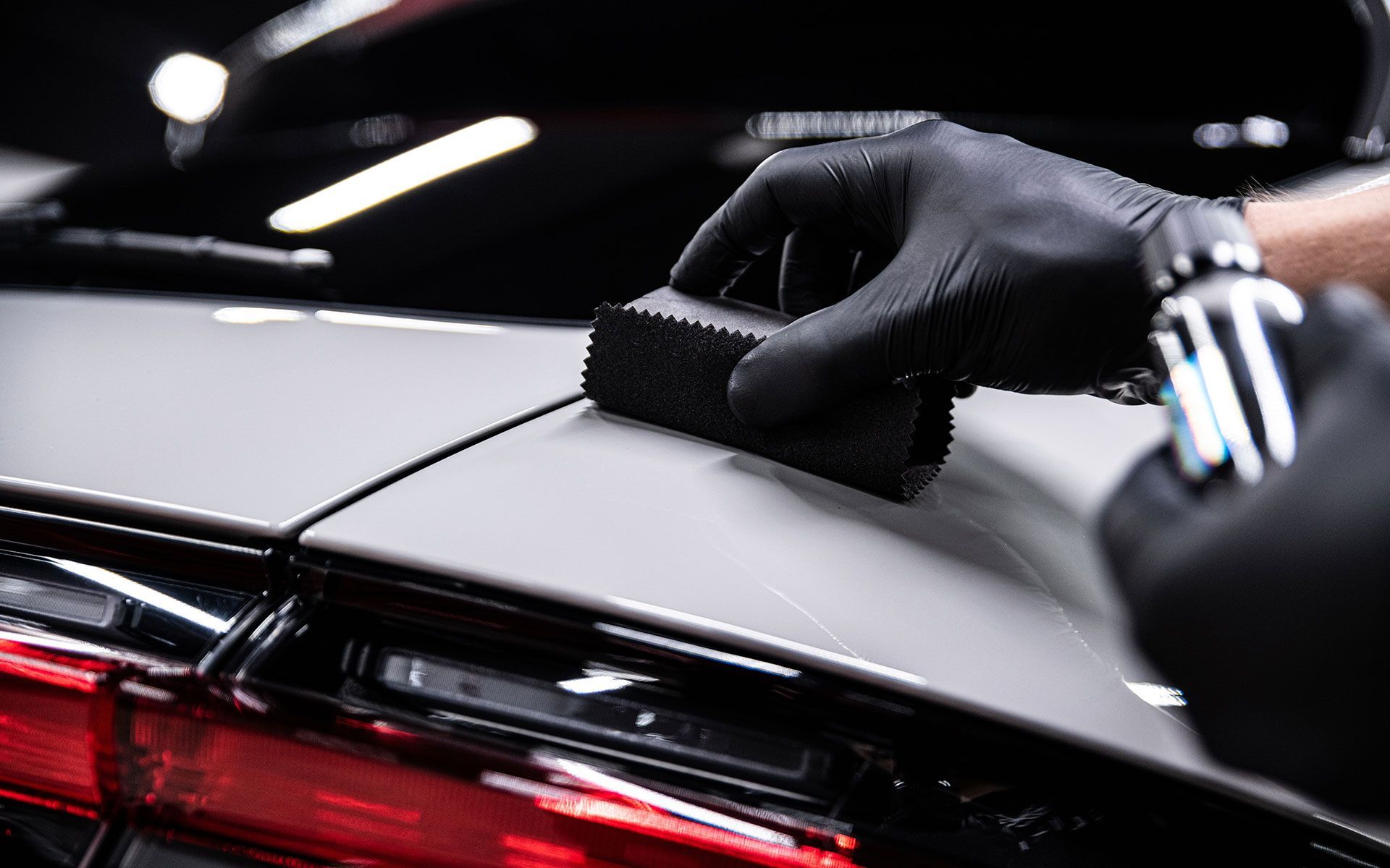 Gloved hand applying ceramic coating on a car's surface with a black applicator sponge and a bottle.