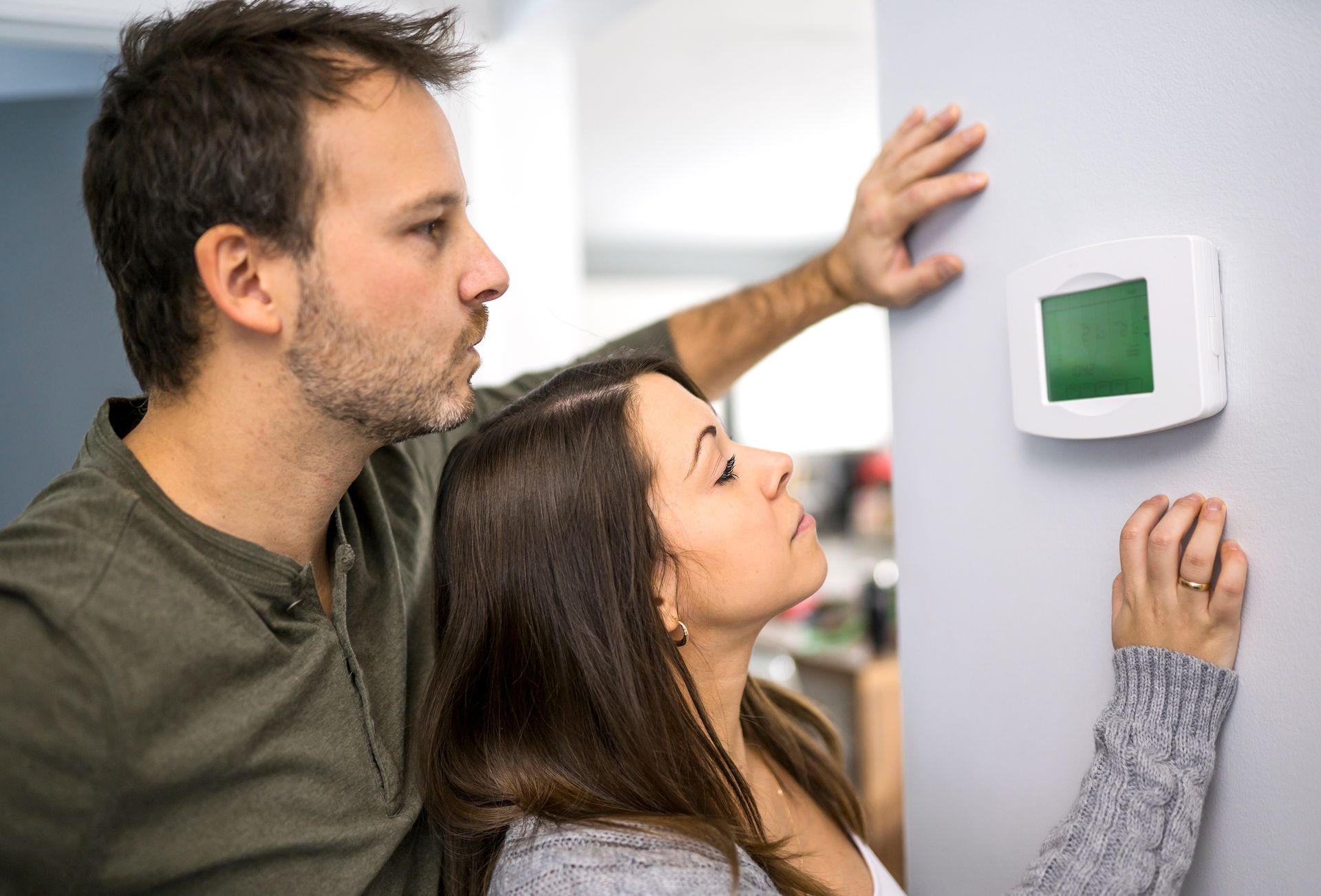 Une personne règle un thermostat numérique blanc fixé au mur tandis qu'une autre personne observe.