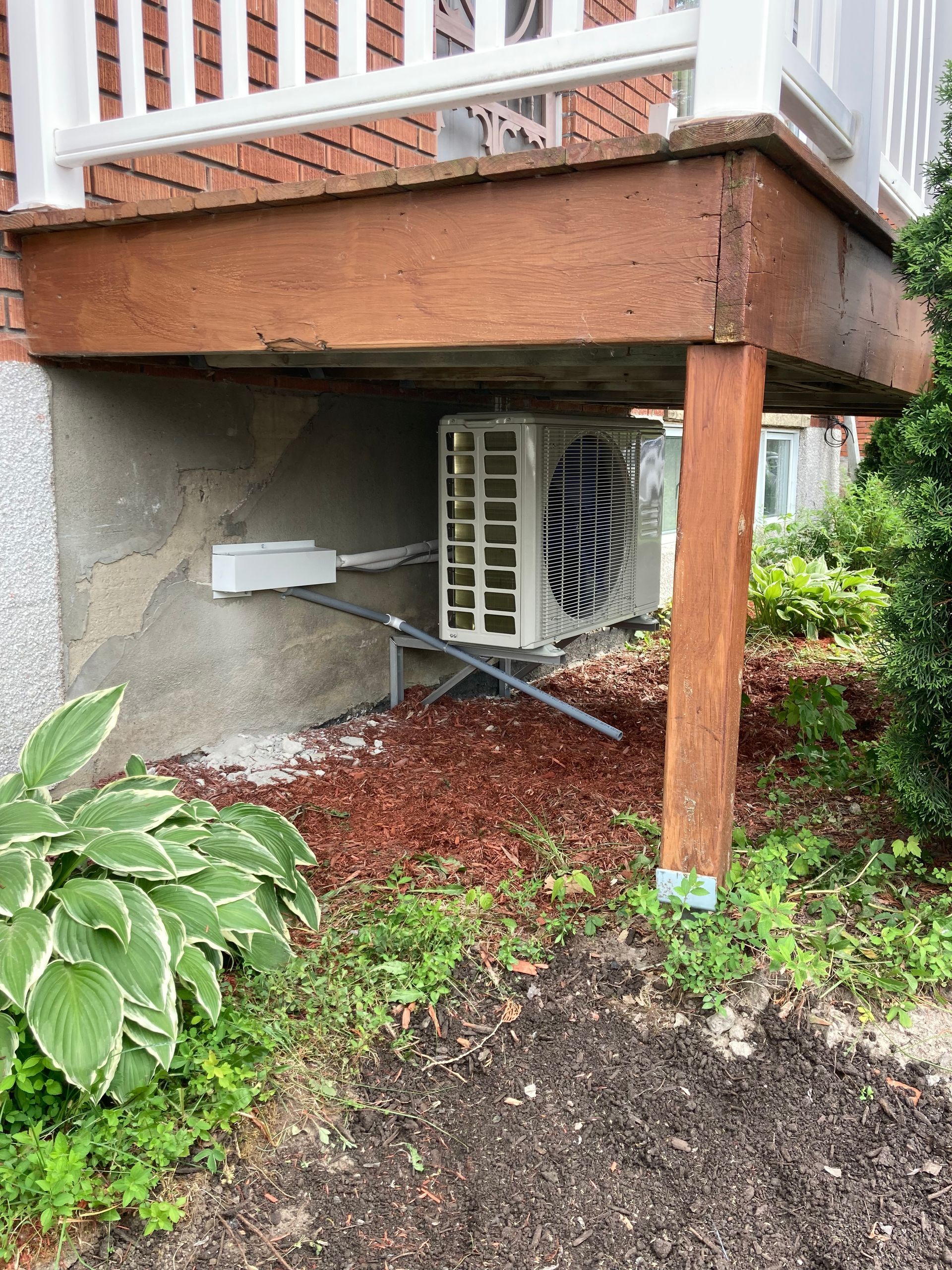 Une unité de pompe à chaleur extérieure est installée sous un porche en bois, posée sur une base de gravier à côté d'un mur de fondation en briques.