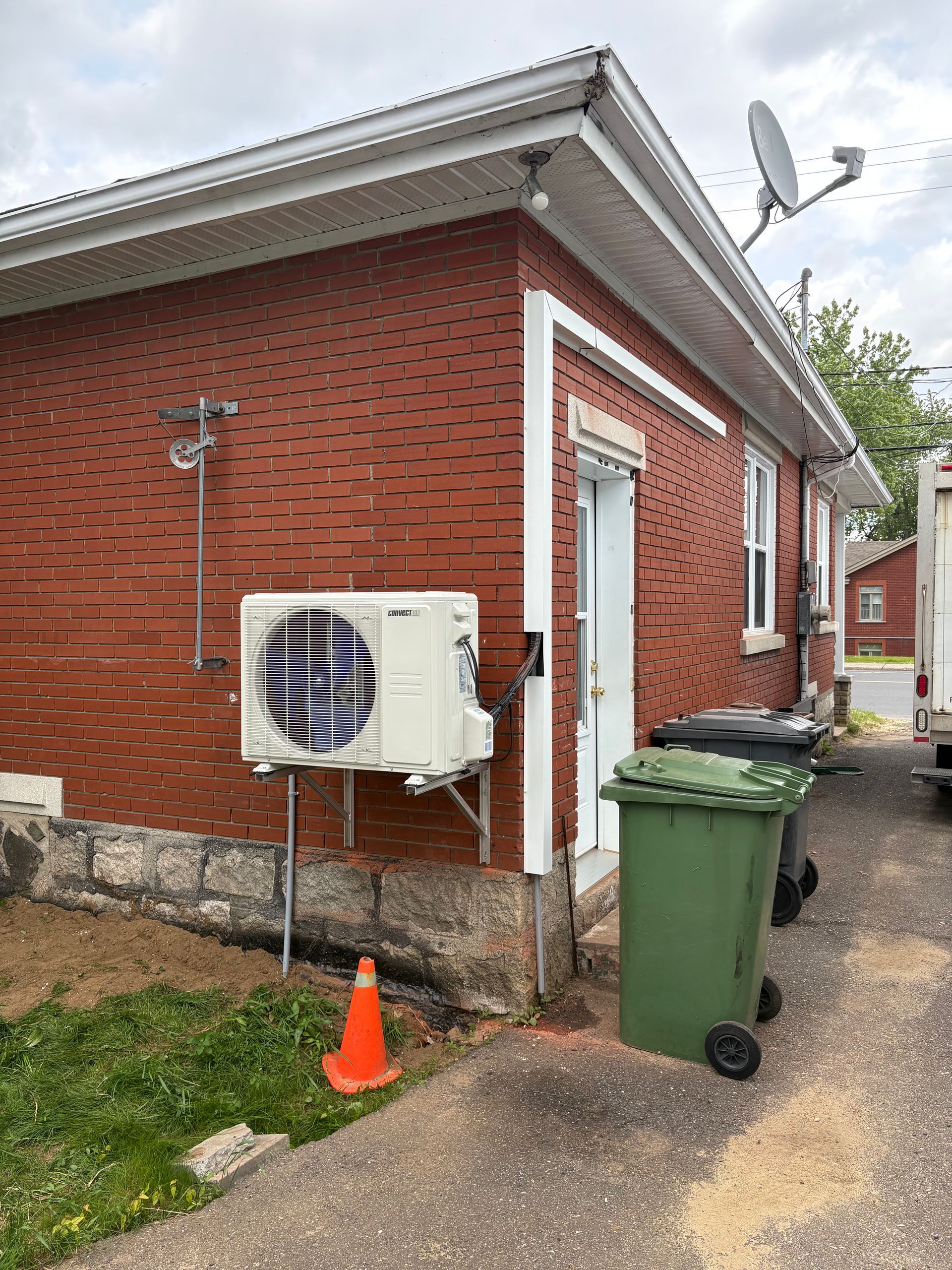 L'extérieur d'une maison en briques rouges présente un climatiseur mural, un cône de signalisation orange et une poubelle verte.