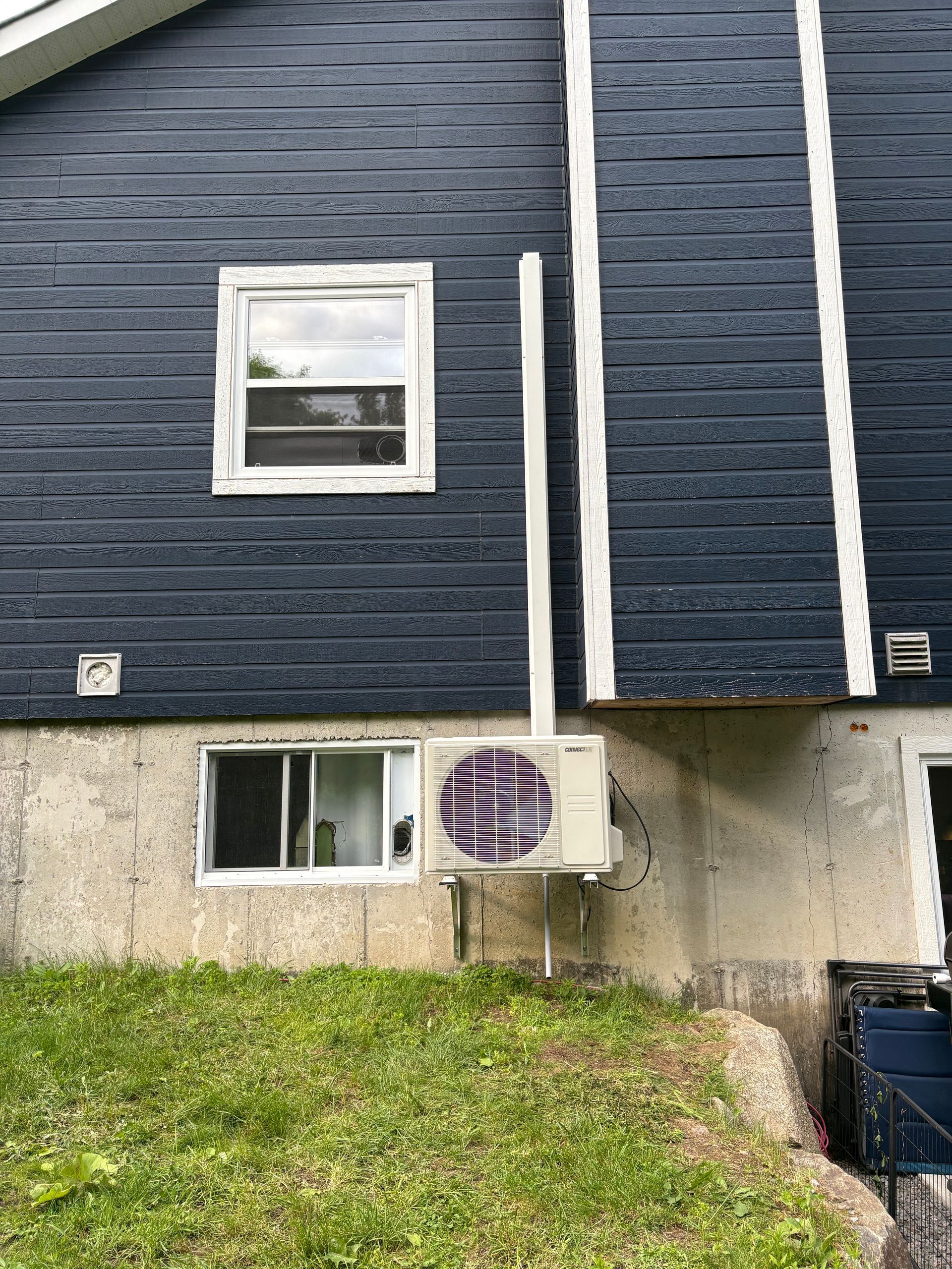 Vue extérieure d'un mur de maison avec des bardeaux foncés, des fondations en béton, des fenêtres et un groupe de condensation extérieur pour la climatisation.