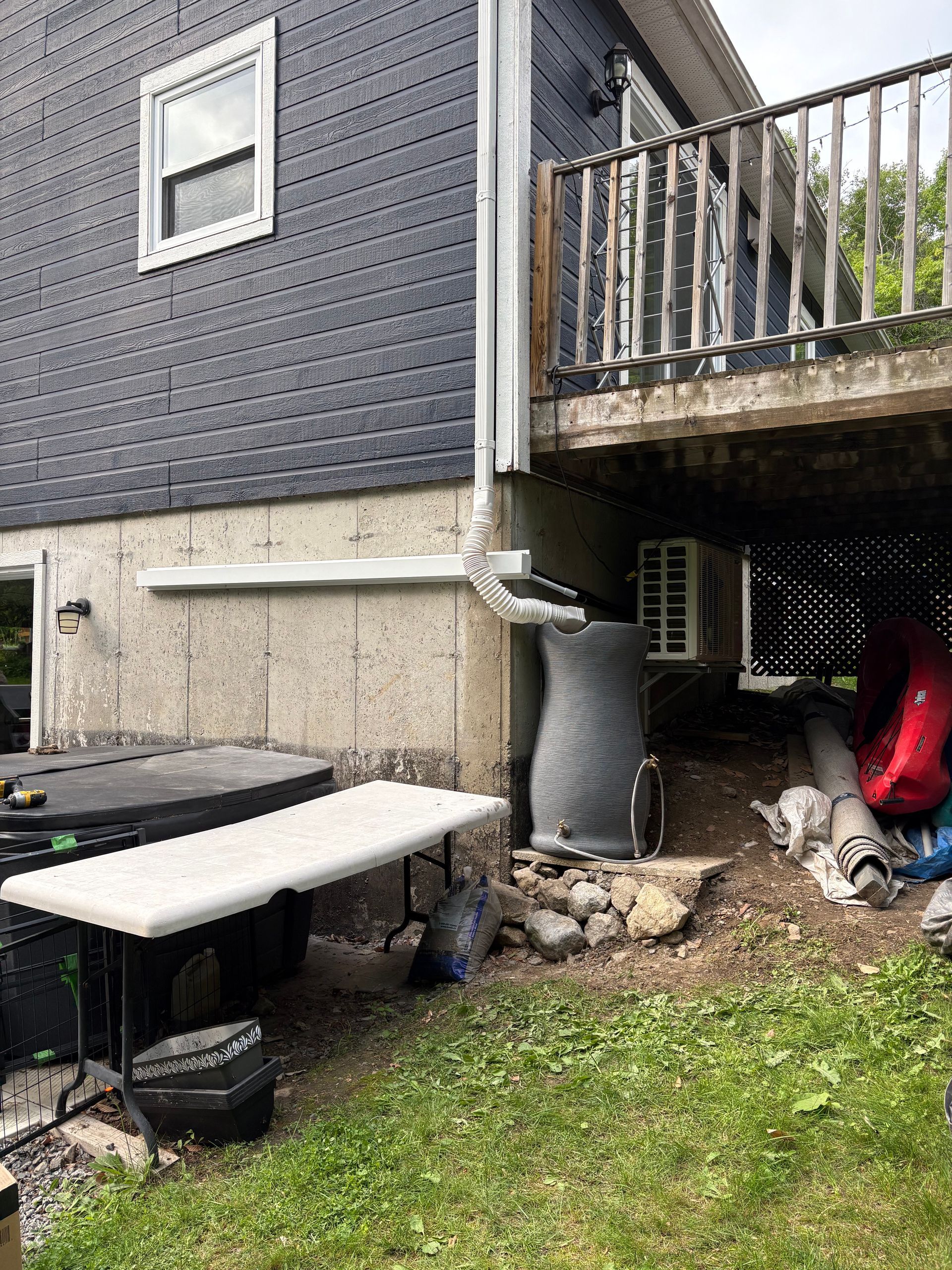 L'extérieur gris d'une maison présente une descente de gouttière redirigée vers un récupérateur d'eau de pluie situé sous une terrasse surélevée.