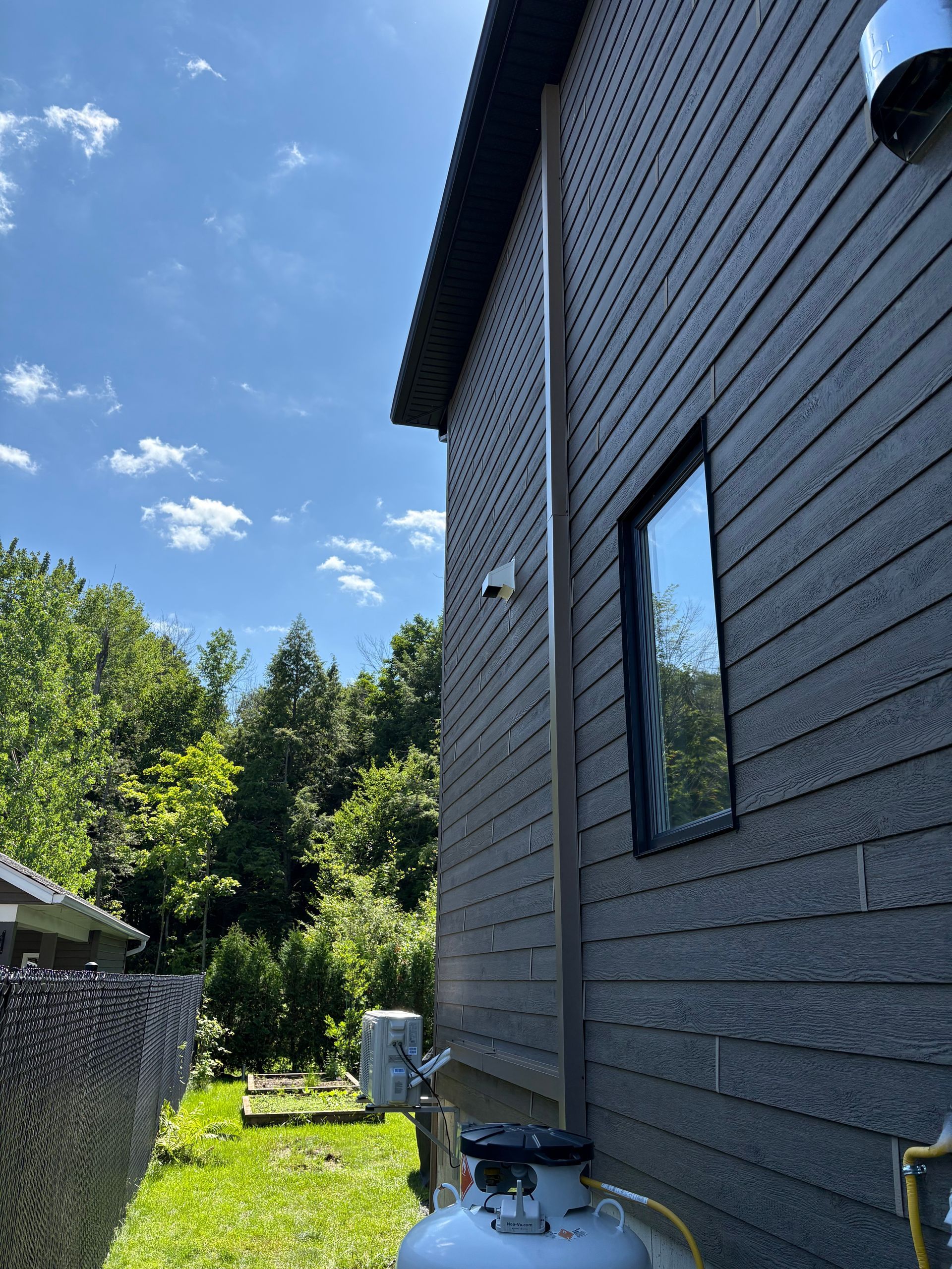Le côté d'une maison gris foncé avec un bardage horizontal, une fenêtre, une descente de gouttière verticale et une bonbonne de propane dans la cour.