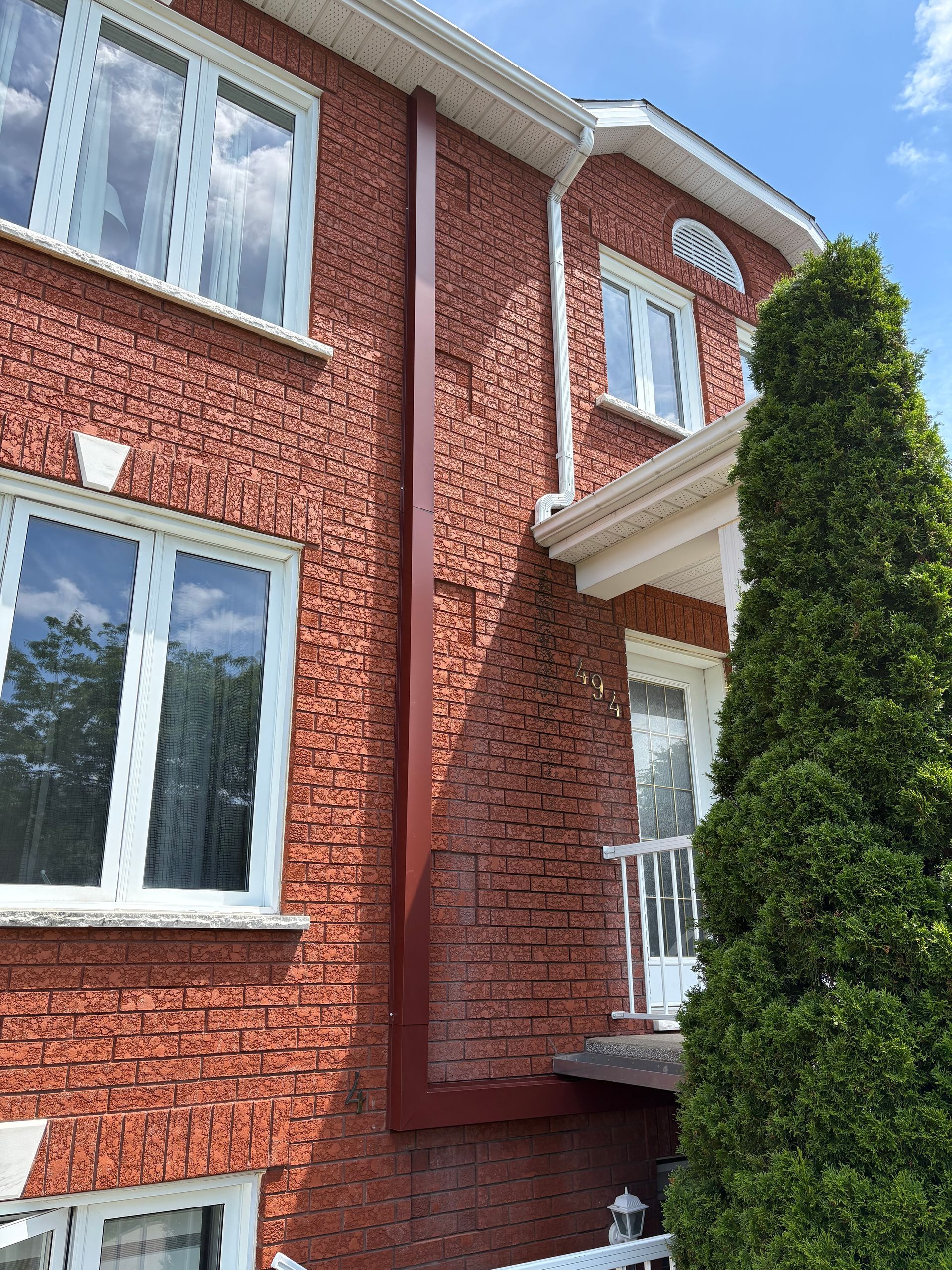 Une maison aux hauts murs de briques rouge foncé, avec des fenêtres à cadre blanc, un balcon blanc et une descente de gouttière proéminente brun foncé.