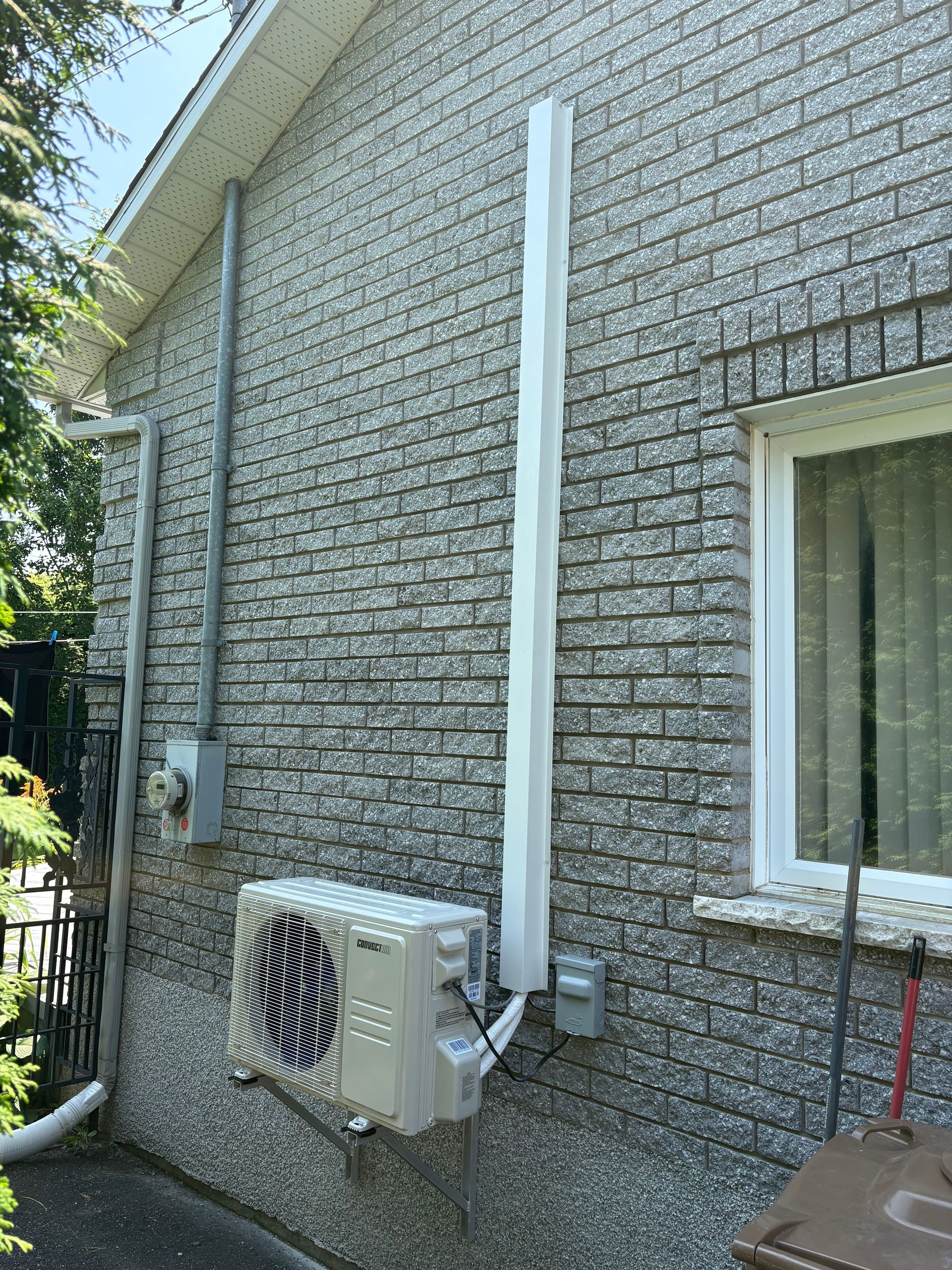 Un climatiseur split extérieur monté sur un mur de briques grises avec un cache-conduit blanc s'étendant vers le haut.