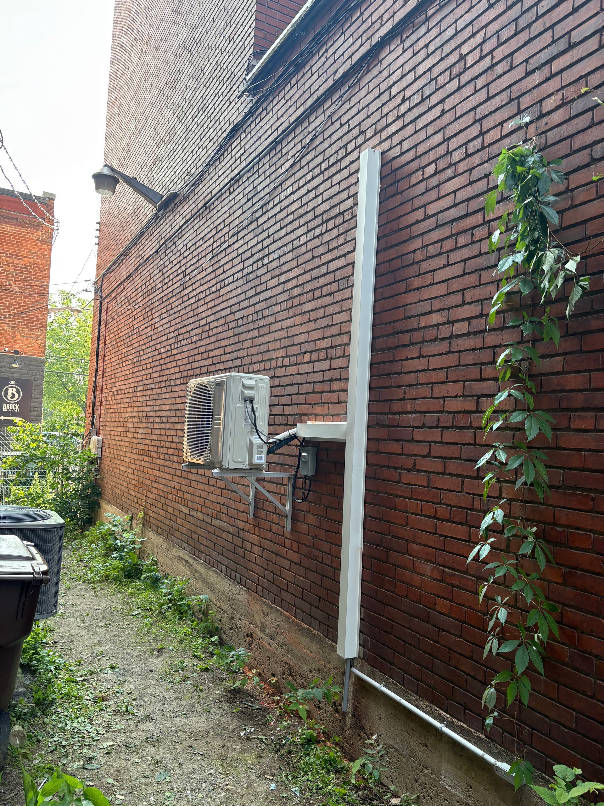 Un climatiseur mural fixé sur un mur extérieur en briques rouges, à côté d'un tuyau blanc vertical et de plantes grimpantes.