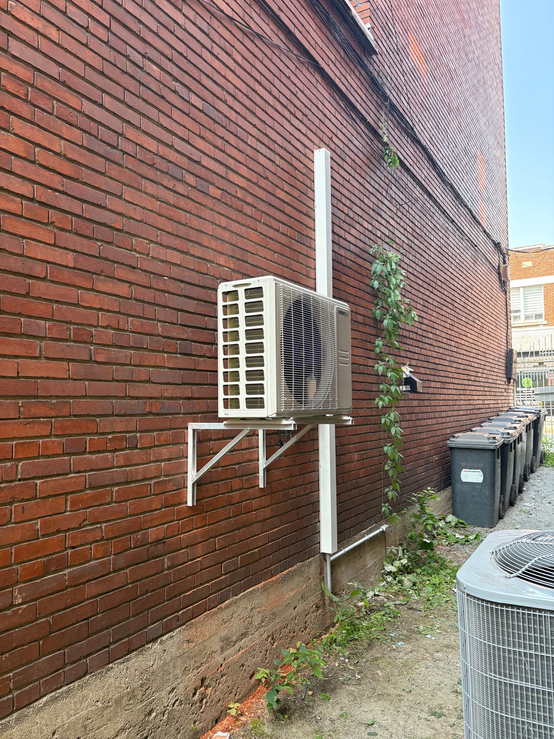 Un climatiseur fixé sur un mur extérieur en briques, au-dessus d'une rangée de poubelles.