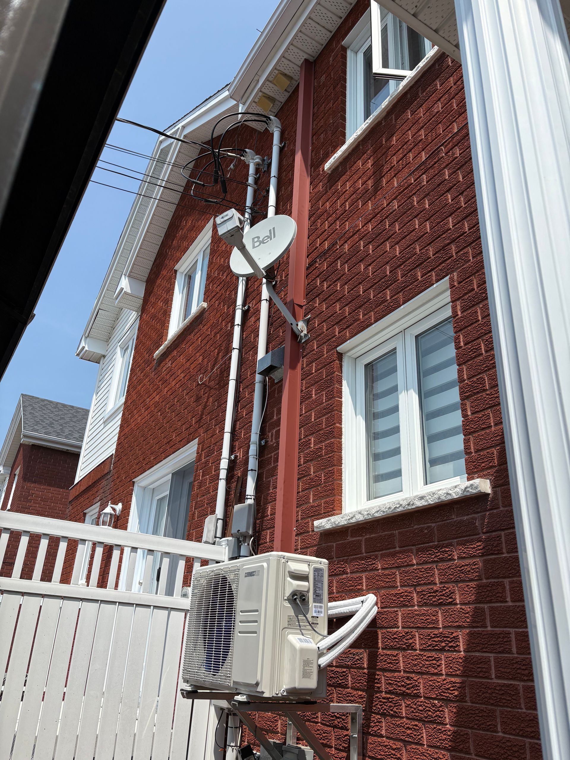Façade en briques rouges d'une maison à deux étages comprenant une antenne parabolique, des canalisations et une unité de climatisation extérieure sur un support.