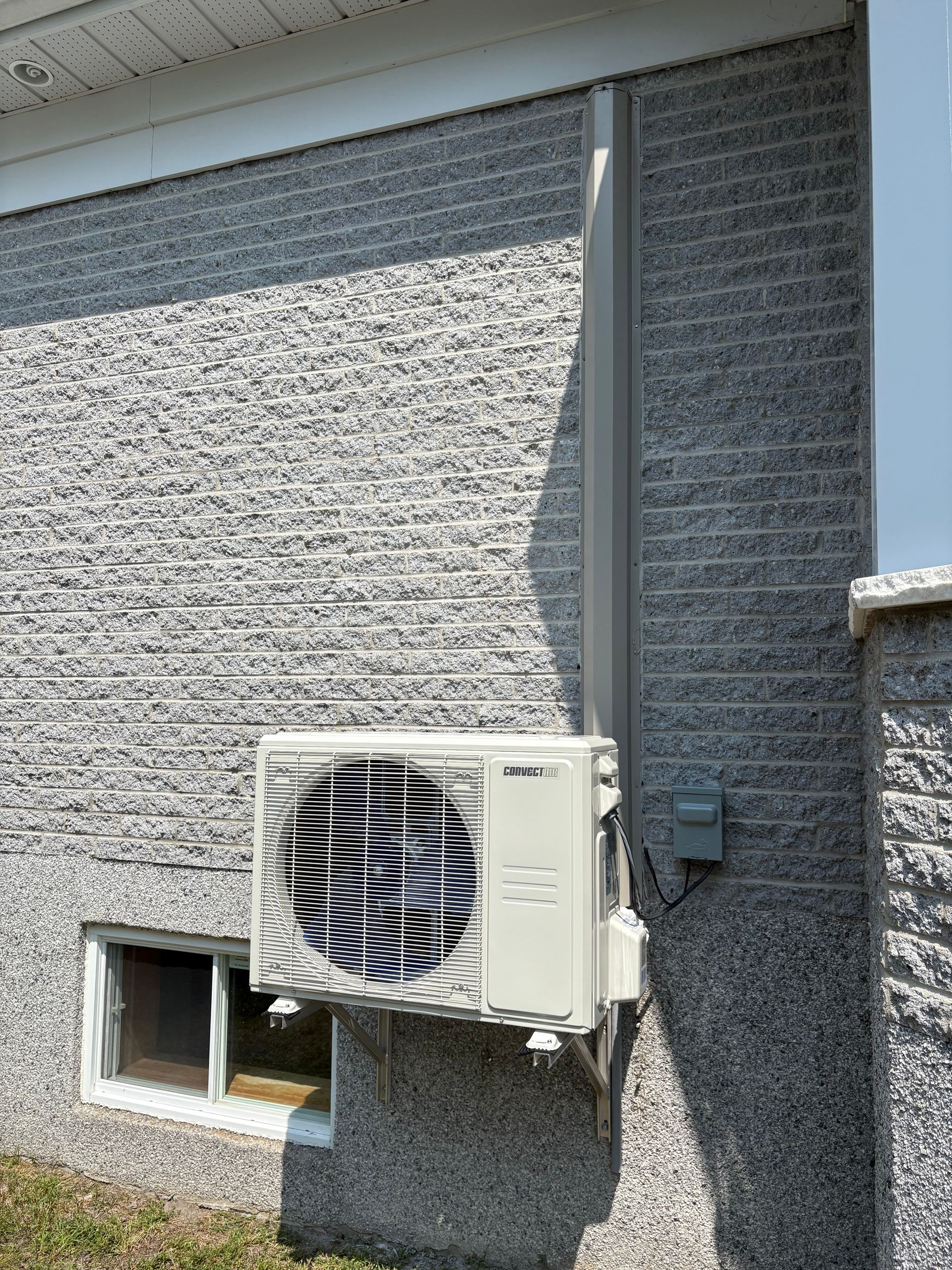 Un condenseur de climatiseur split beige, monté sur un mur extérieur en briques grises texturées, à côté d'une petite fenêtre.