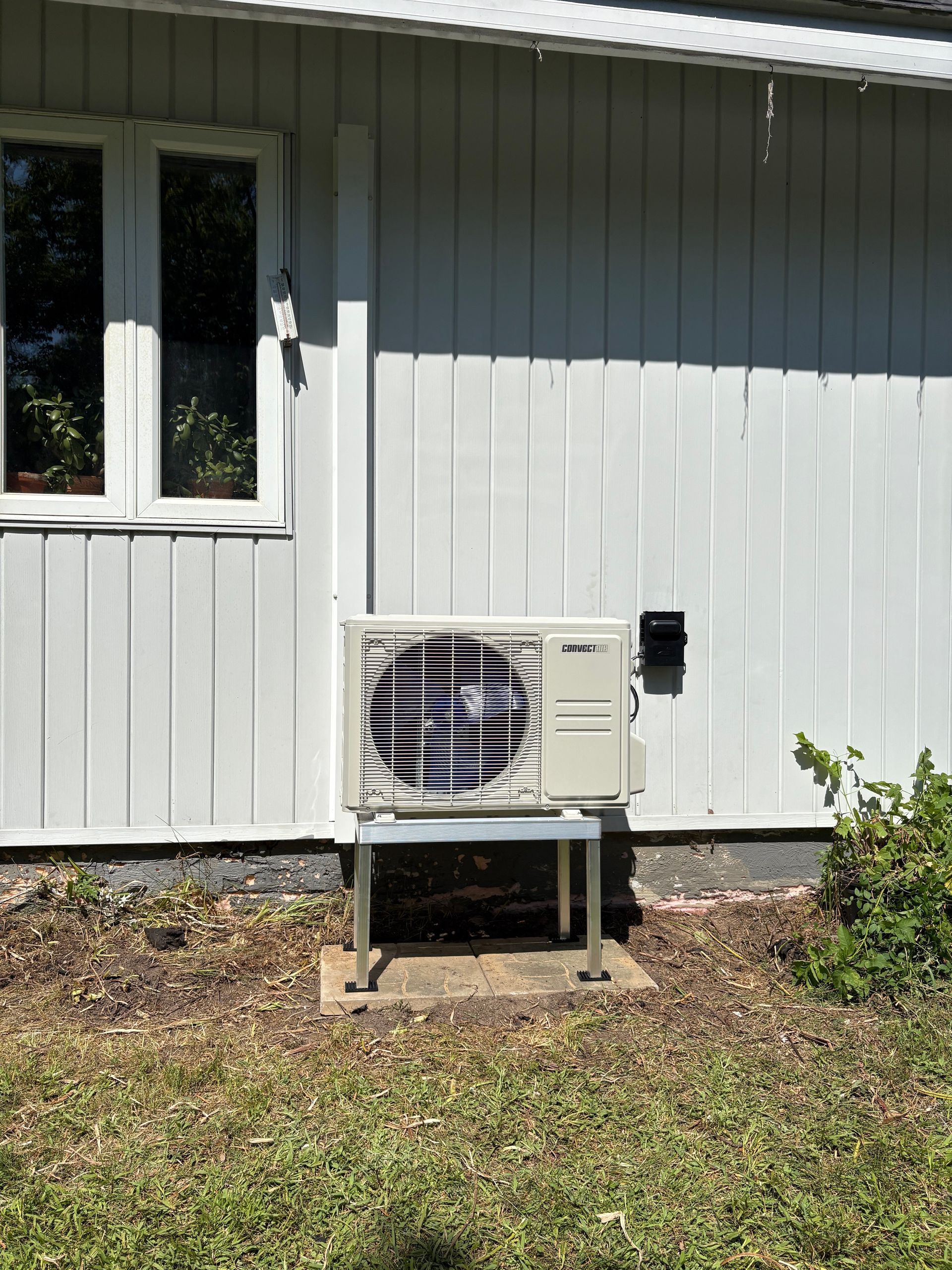 Un climatiseur extérieur beige repose sur un support métallique, adossé au bardage blanc à panneaux verticaux d'une maison.
