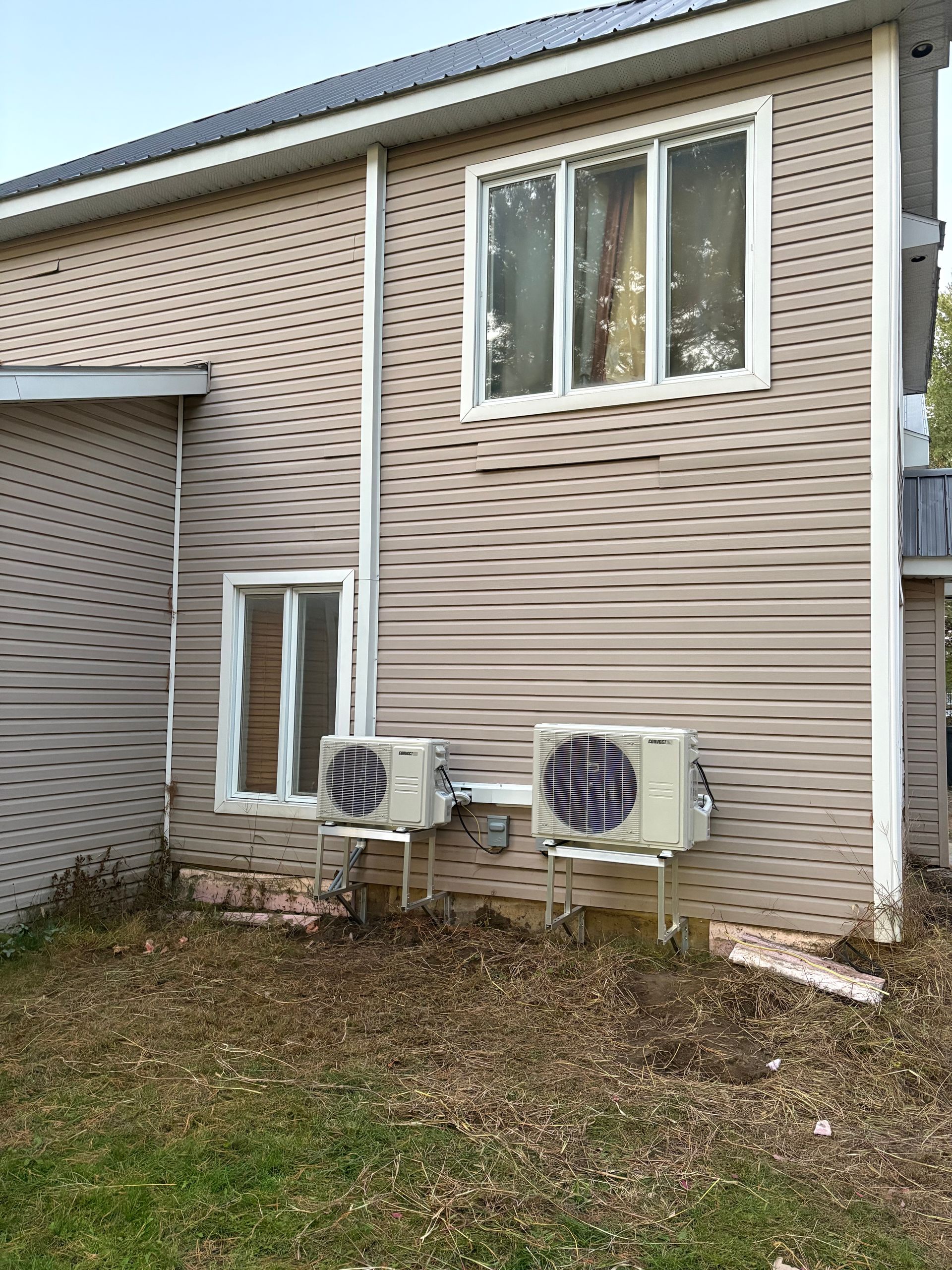 Deux unités de climatisation extérieures montées sur le revêtement beige d'une maison à deux étages avec deux fenêtres et un toit en métal.